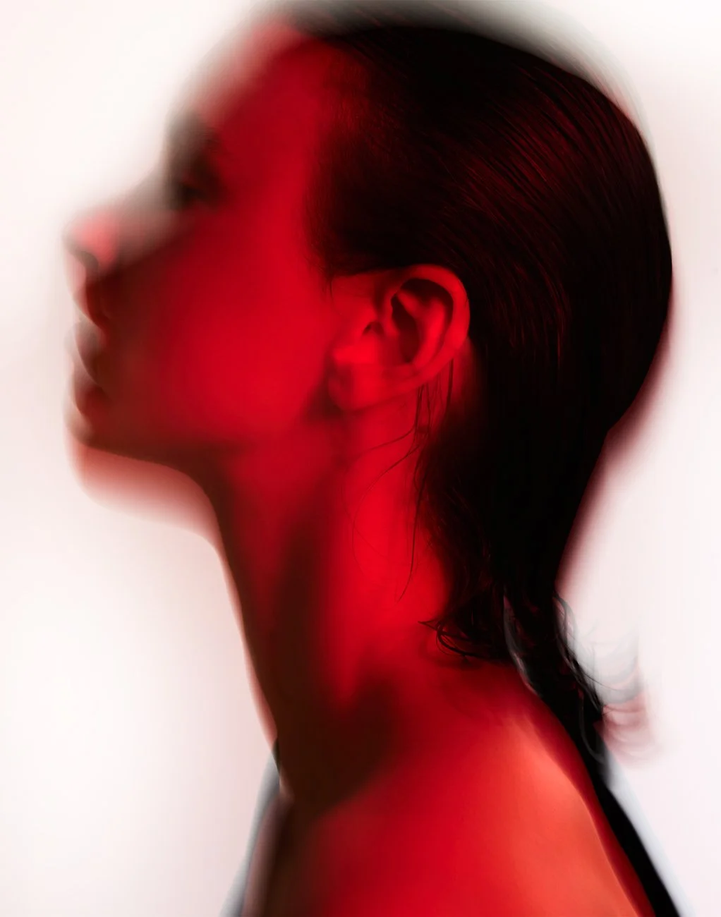 A close-up side profile of a woman with dark hair, bathed in red light, creating a blurred and artistic effect.