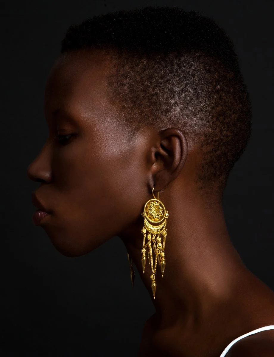 Side profile of a woman with short, textured haircut, wearing large gold earrings with intricate designs and dangling elements, set against a dark background.