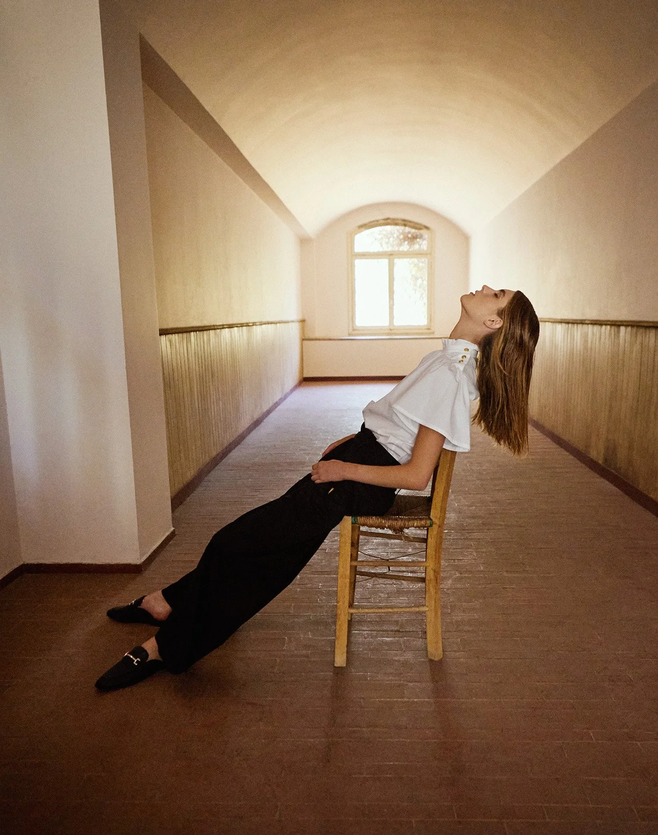 A woman with long brown hair sitting backward on a wooden chair in a hallway, with her head tilted back and eyes closed, wearing a white blouse and black pants.