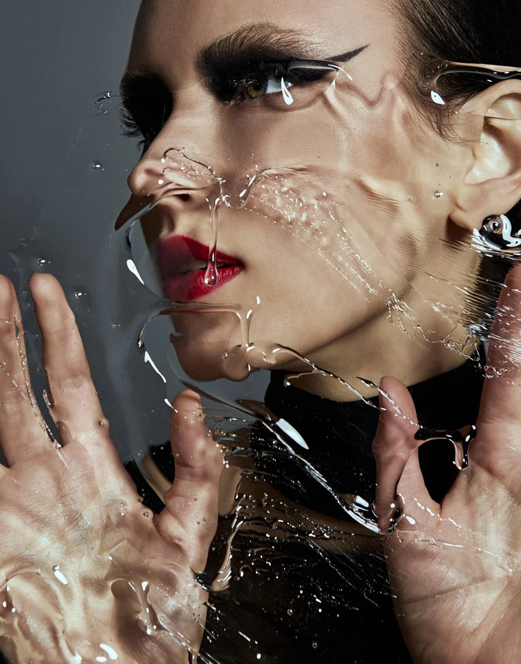 A woman with makeup behind a glass surface covered in water droplets, touching the glass with both hands.