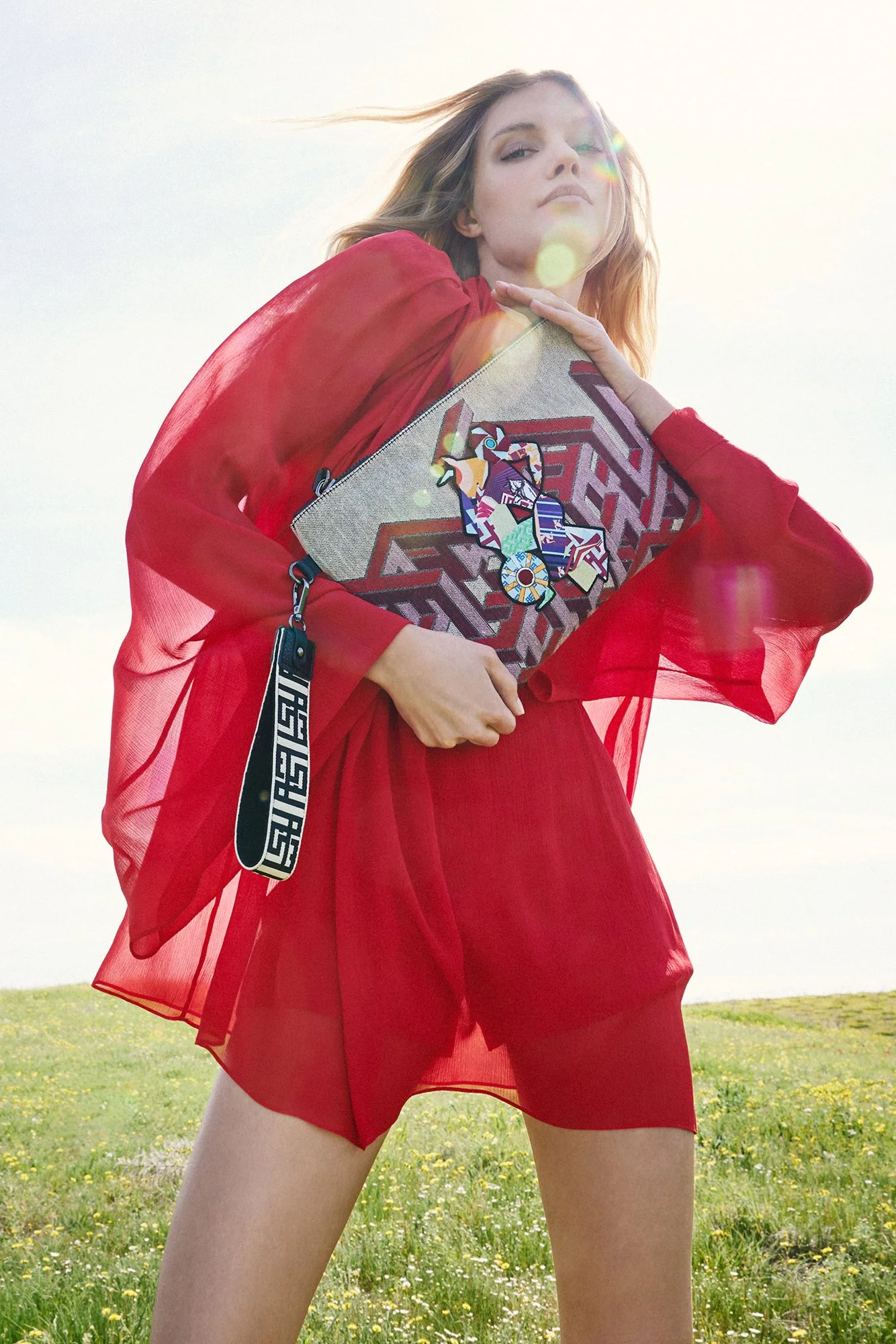 A woman in a red sheer dress holding a handbag with colorful designs, standing outdoors in a grassy field with yellow flowers, sunlight creating lens flares.