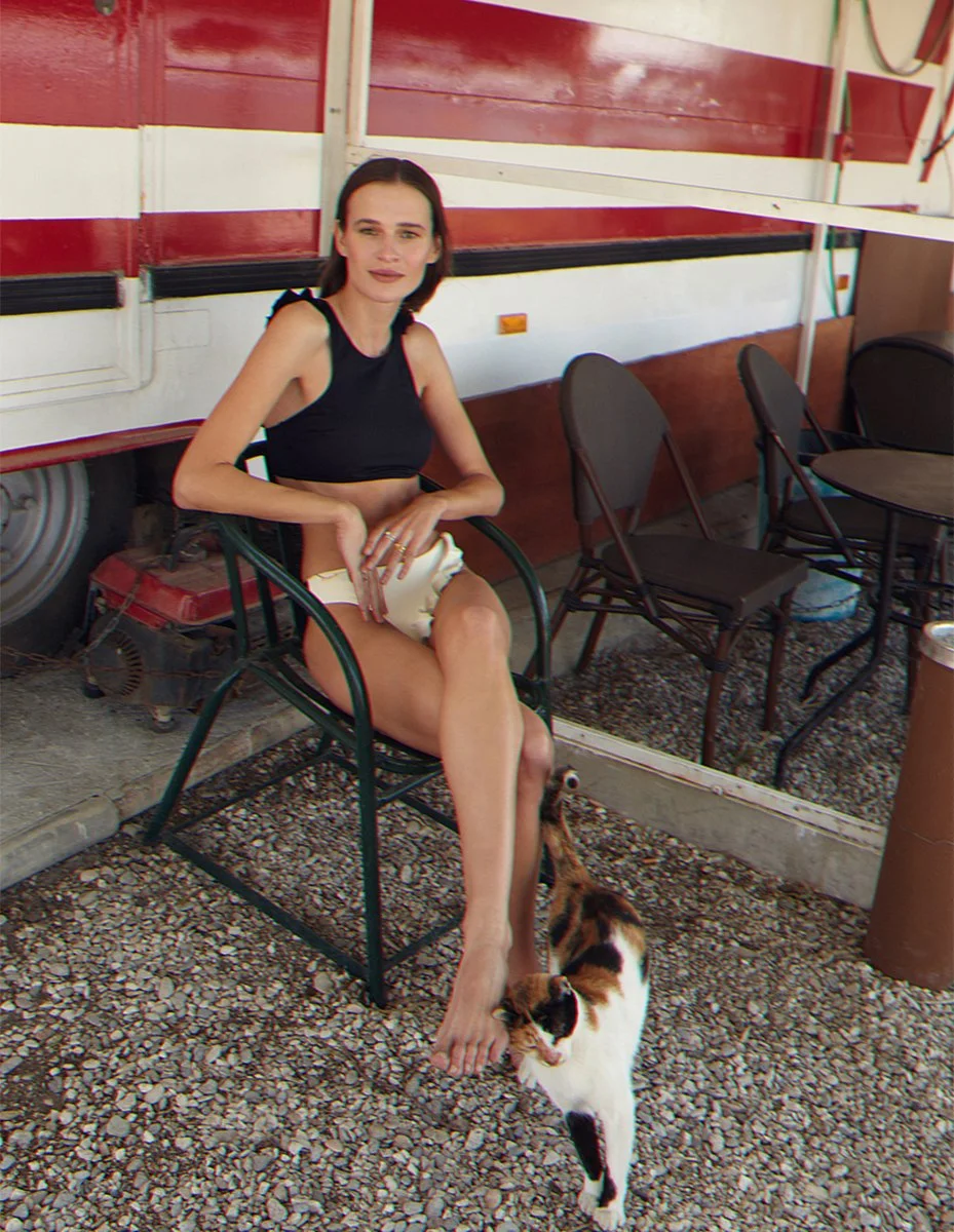 A woman with dark hair, wearing a black sleeveless top and beige shorts, seated outdoors on a black chair, with a calico cat walking by her feet. The background features red and white painted wooden walls, outdoor chairs, and a gravel ground.