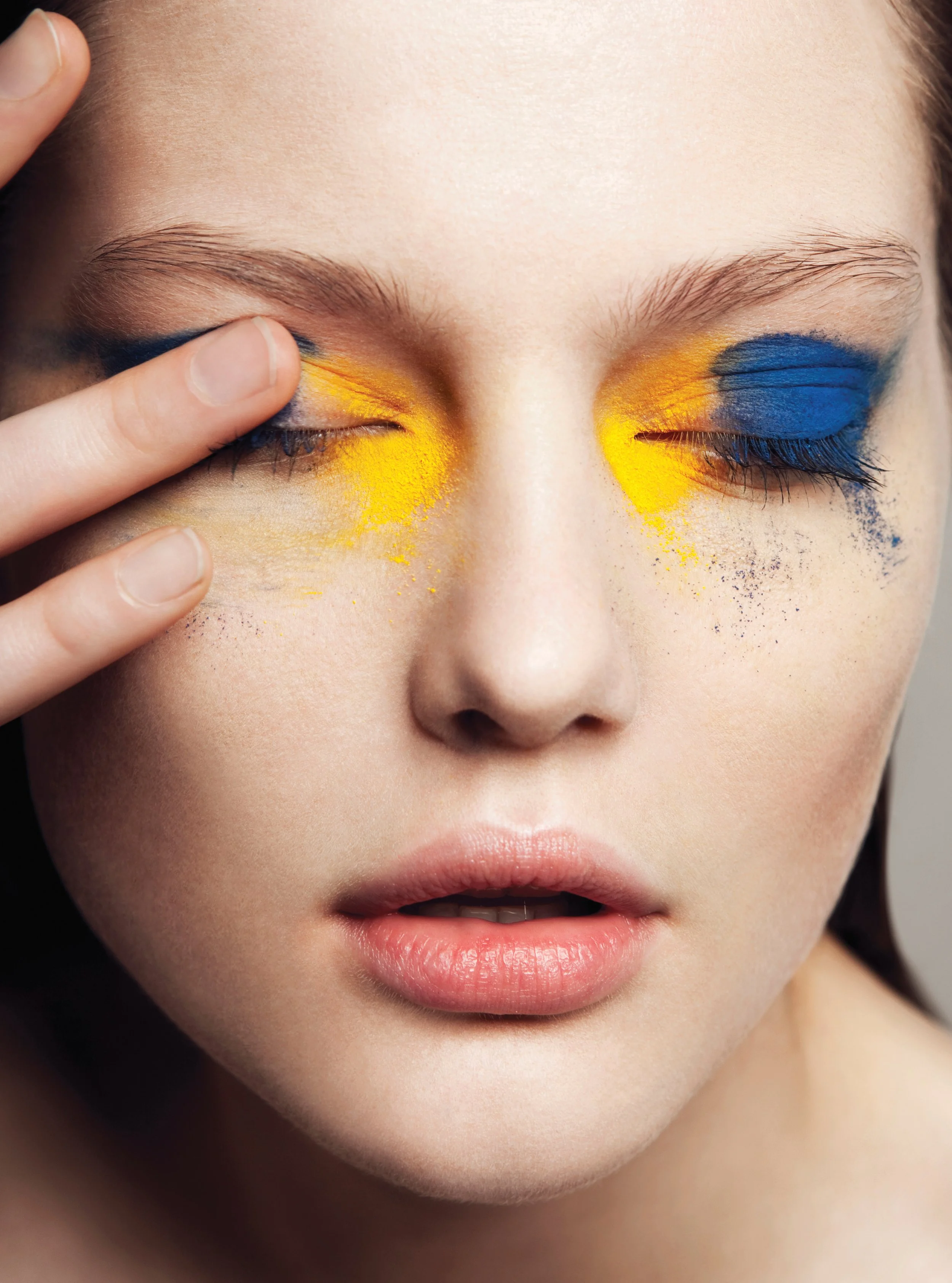 Close-up of a woman's face with eyes closed, showing vibrant yellow and blue eyeshadow, with yellow powder on cheeks.