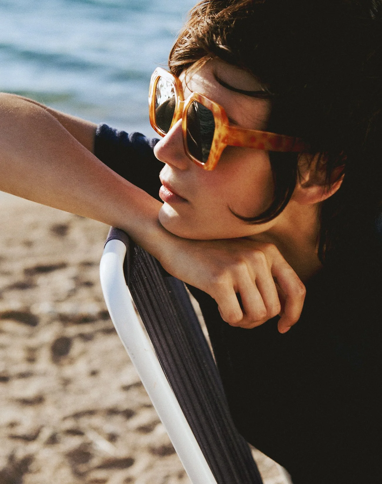 Close-up of a woman wearing tortoiseshell sunglasses, resting her elbow on a chair on the beach, with the ocean in the background.