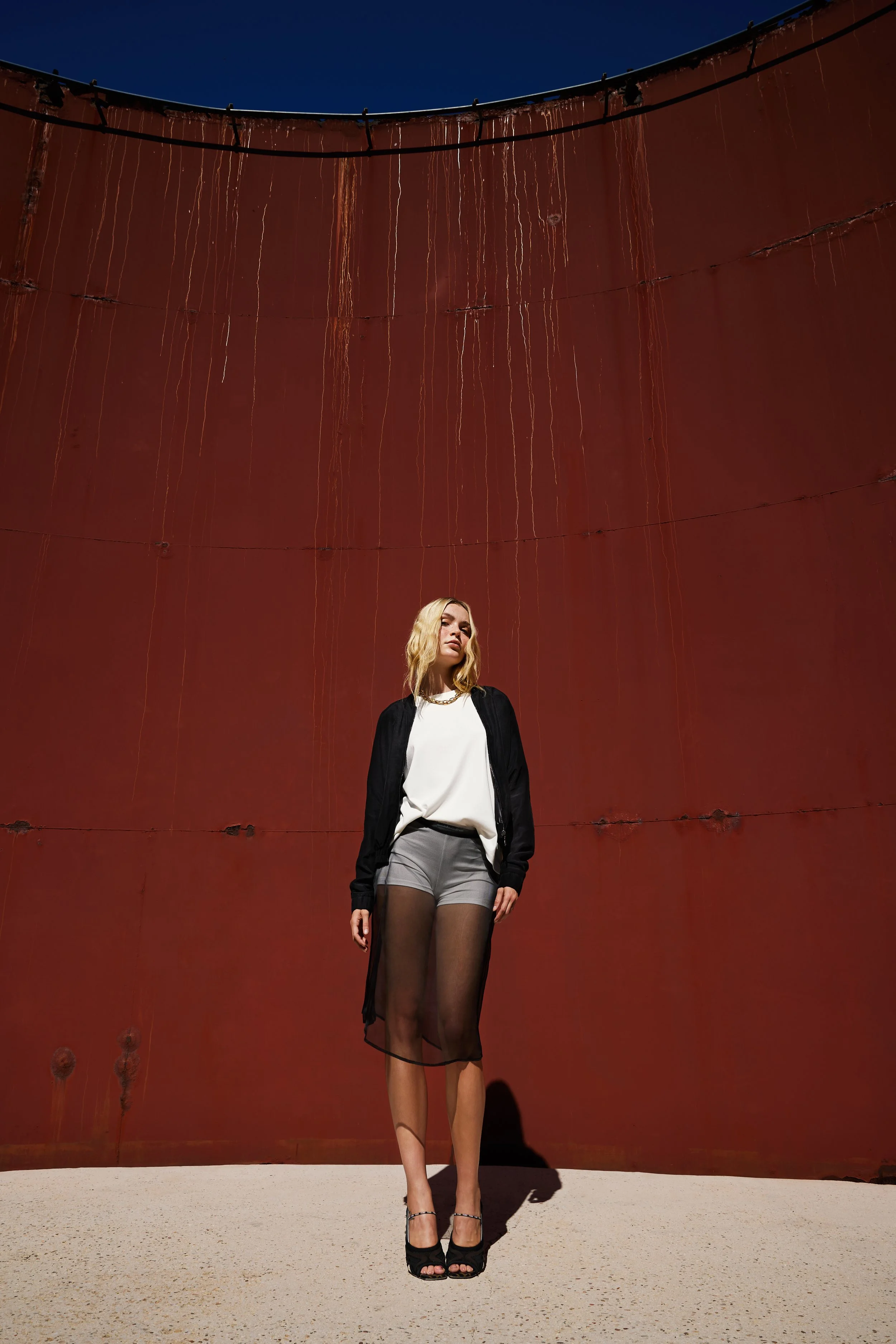 A young woman stands in front of a large, rusty, curved wall with streaks of rust and peeling paint, under a clear blue sky. She is wearing a white top, black jacket, sheer black skirt, and black high heels, with blonde hair and a serious expression.