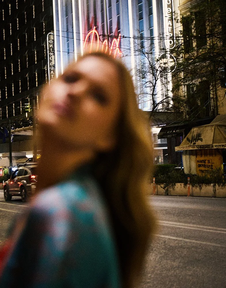 A blurred woman with long hair stands on a city street at night, with neon lights and tall buildings in the background.