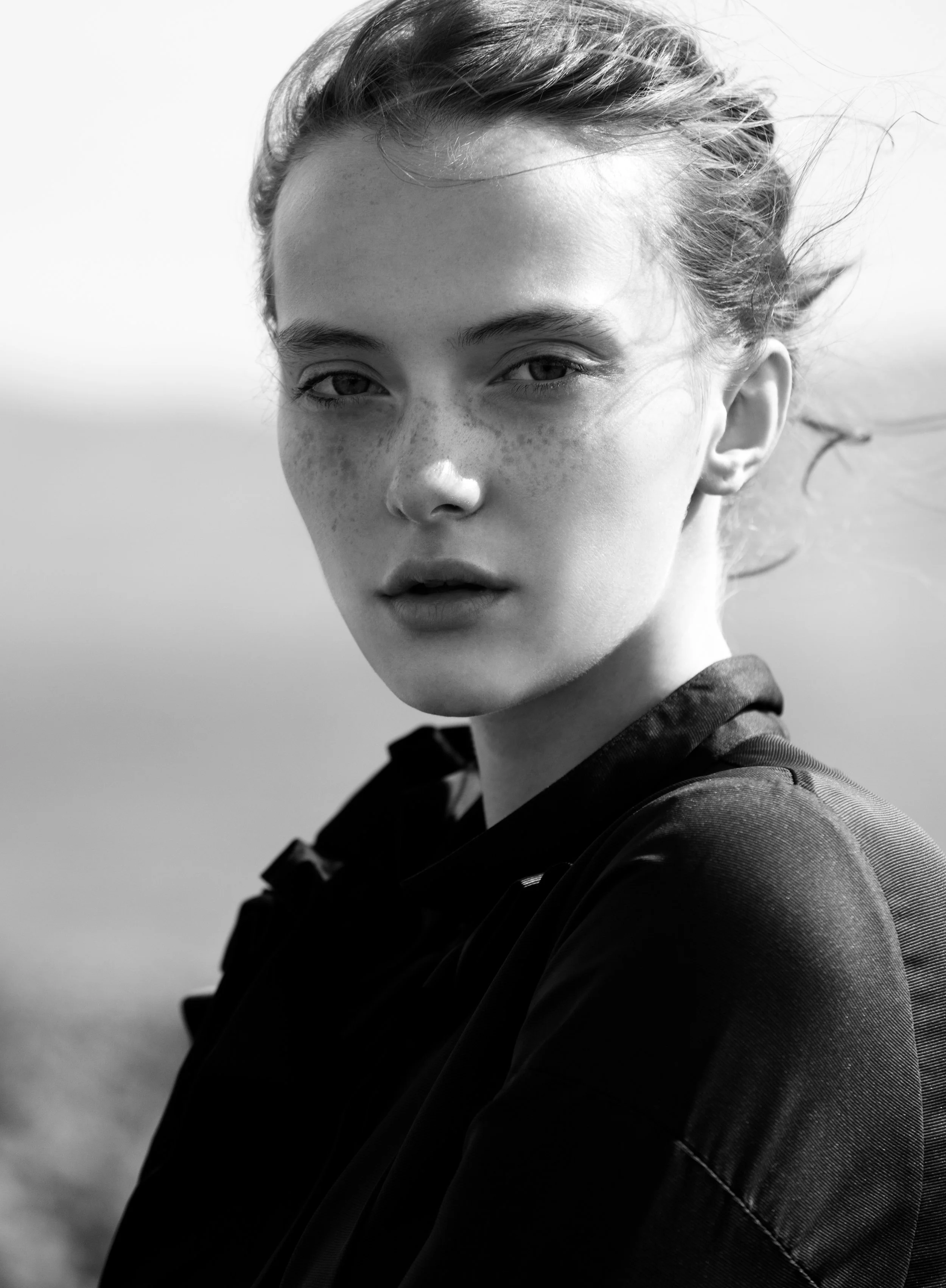 Black and white close-up portrait of a young woman with short hair, freckles, and light-colored eyes, looking directly at the camera with a neutral expression, wearing a dark top.