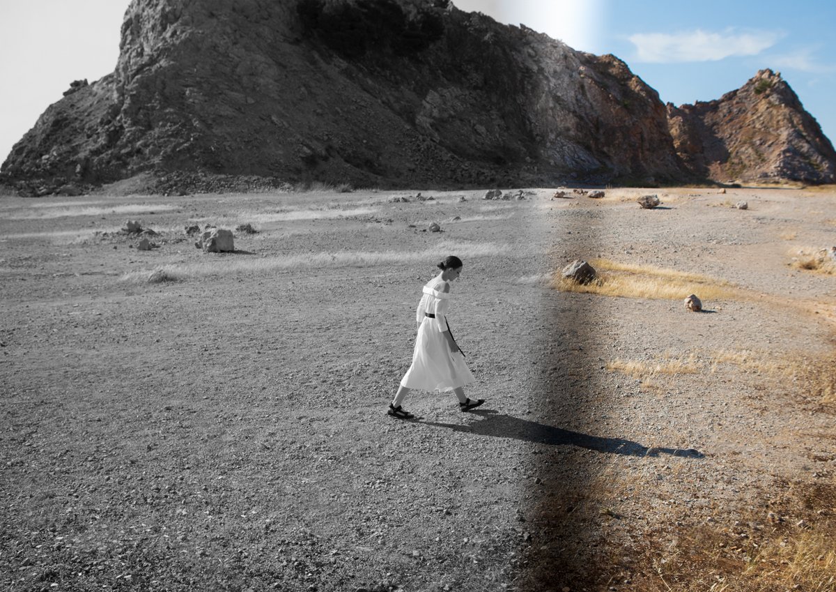 A woman in a white dress and black sandals walking on a dry, rocky landscape with mountains in the background, with part of the image in black and white and part in color.