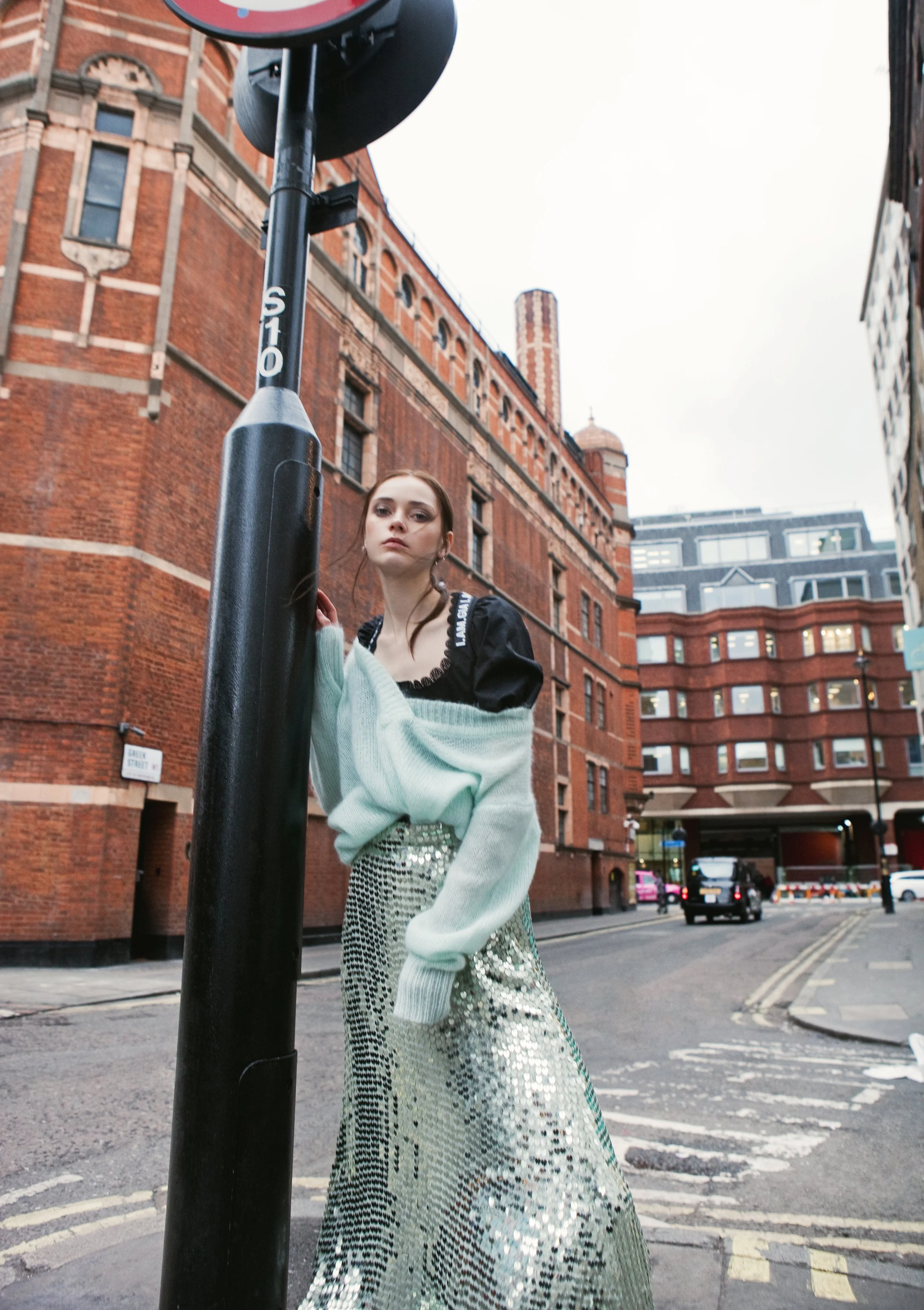 A young woman with long dark hair standing on a city street near a black pole, with a large red-brick building in the background. She is wearing a black top underneath a light-colored oversized sweater and a long, shiny, sequined skirt. The sky is ov
