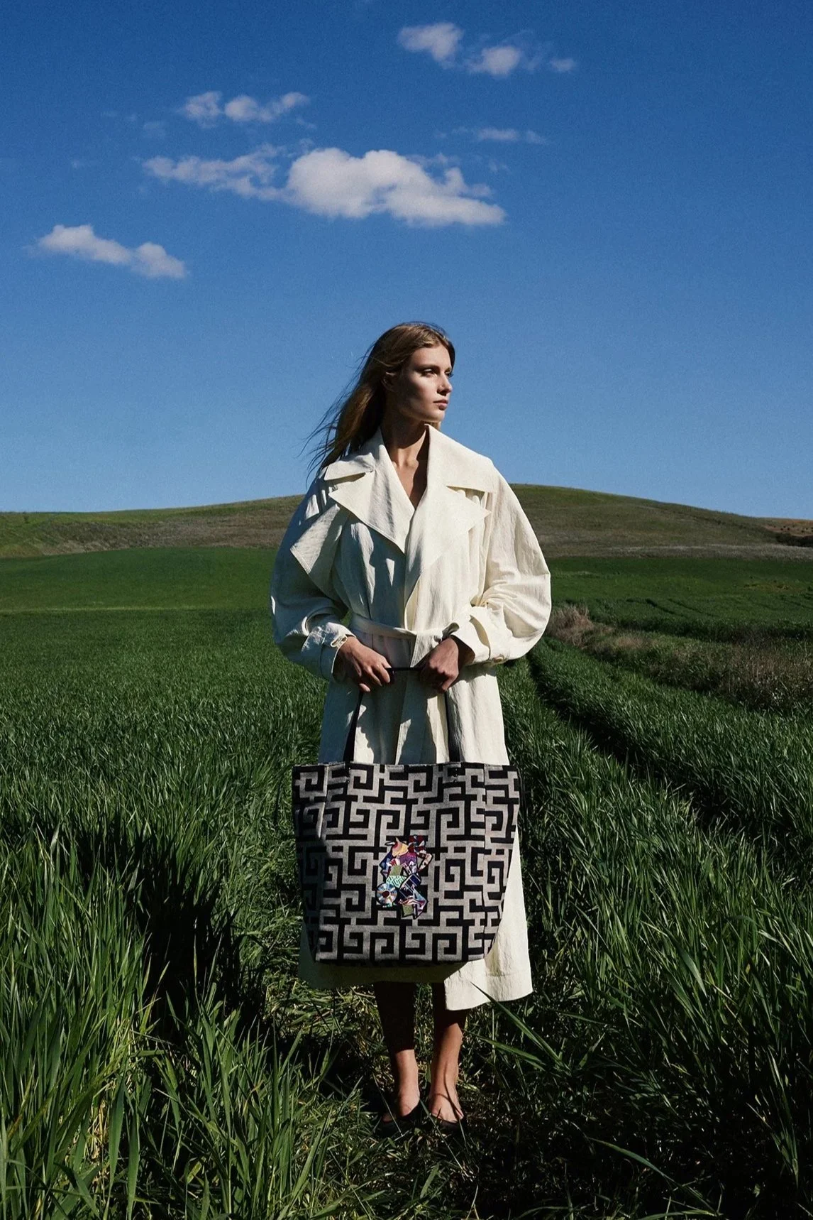 A woman standing in a green field under a blue sky with a few clouds, holding a patterned bag with a colorful design on it.