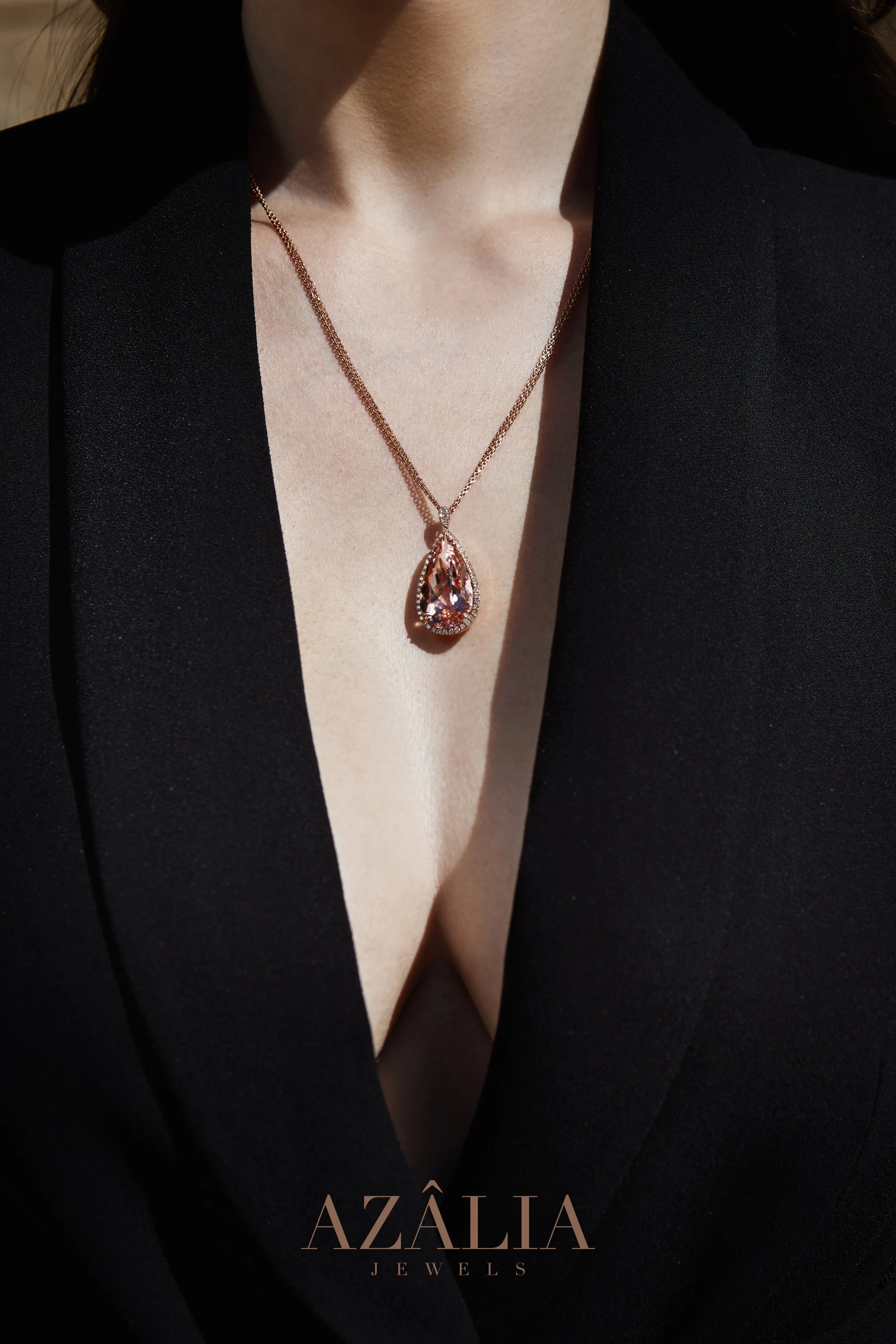 Close-up of a woman wearing a black blazer and a gold necklace with a large, teardrop-shaped pendant. The image includes the text 'AZÁLIA JEWELS' at the bottom.