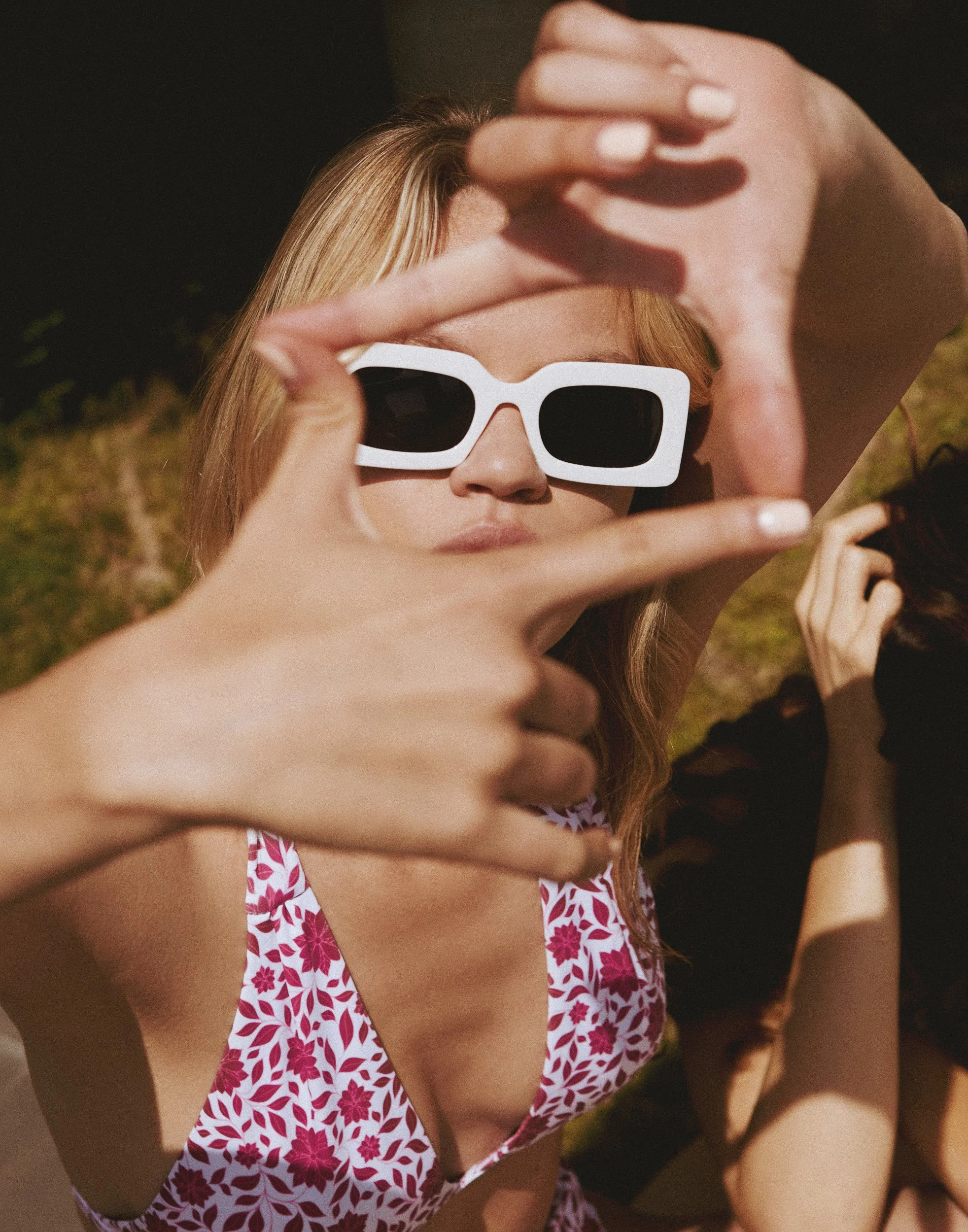 A woman with blonde hair wearing white sunglasses and a pink and white floral swimsuit making a heart shape with her hands outdoors.