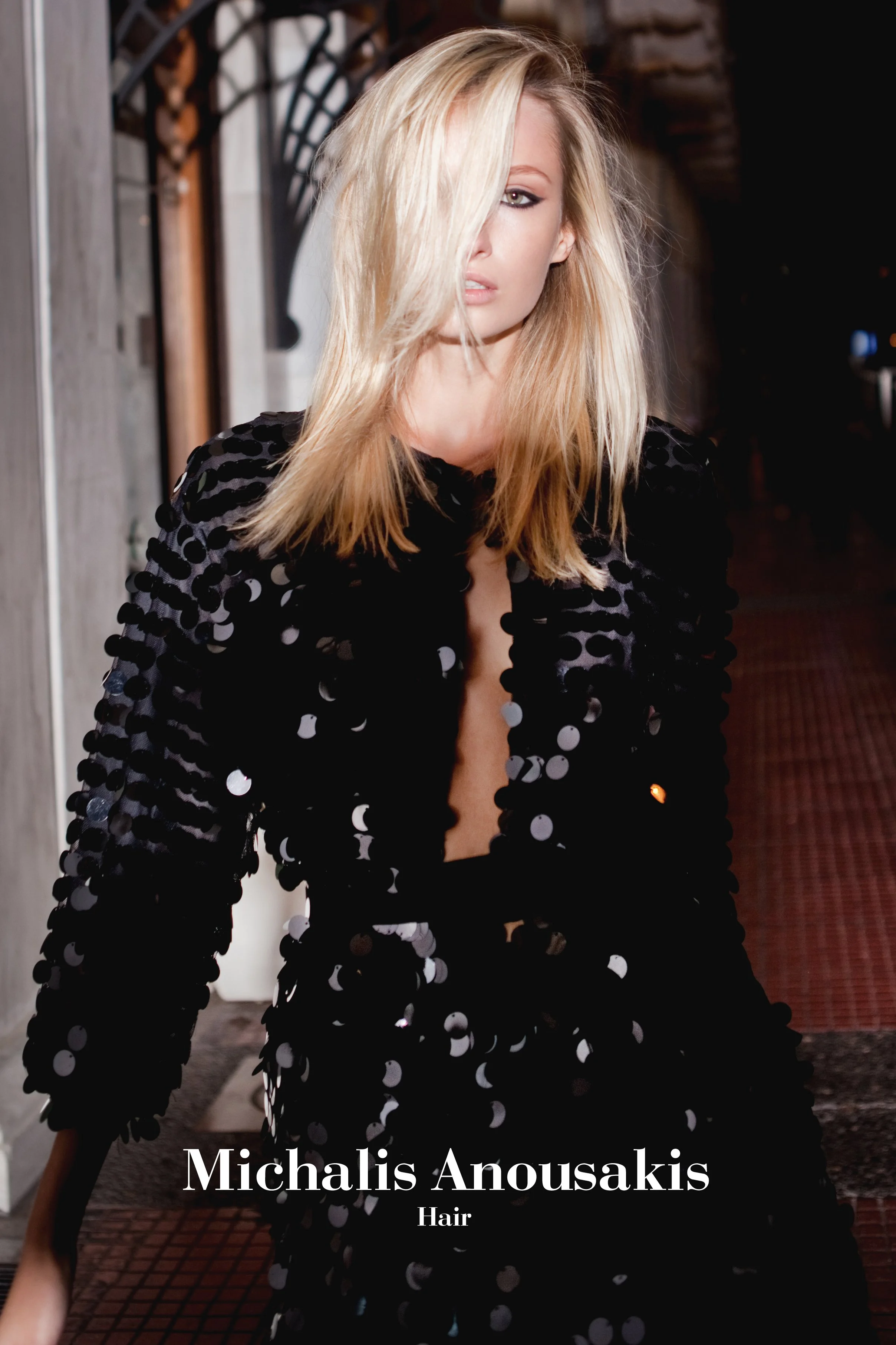 A woman with blonde hair partially covered face, wearing a black jacket with shiny circular embellishments, standing in front of a background with wood and metal structures and a dark corridor. Text overlay reads: 'Michalis Anousakis Hair'.