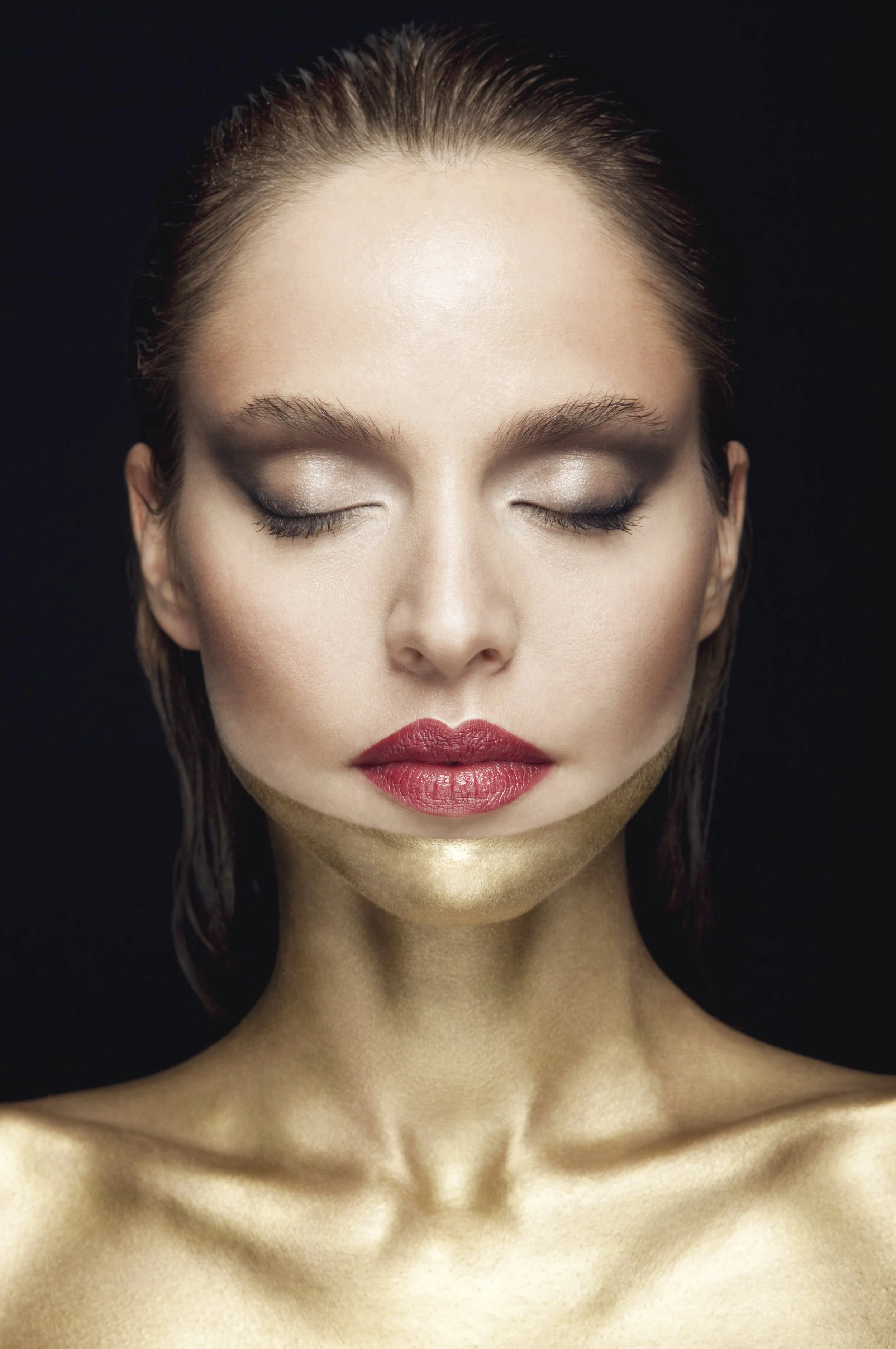 A close-up of a woman's face with her eyes closed, showing her makeup and gold painted neck, against a black background.