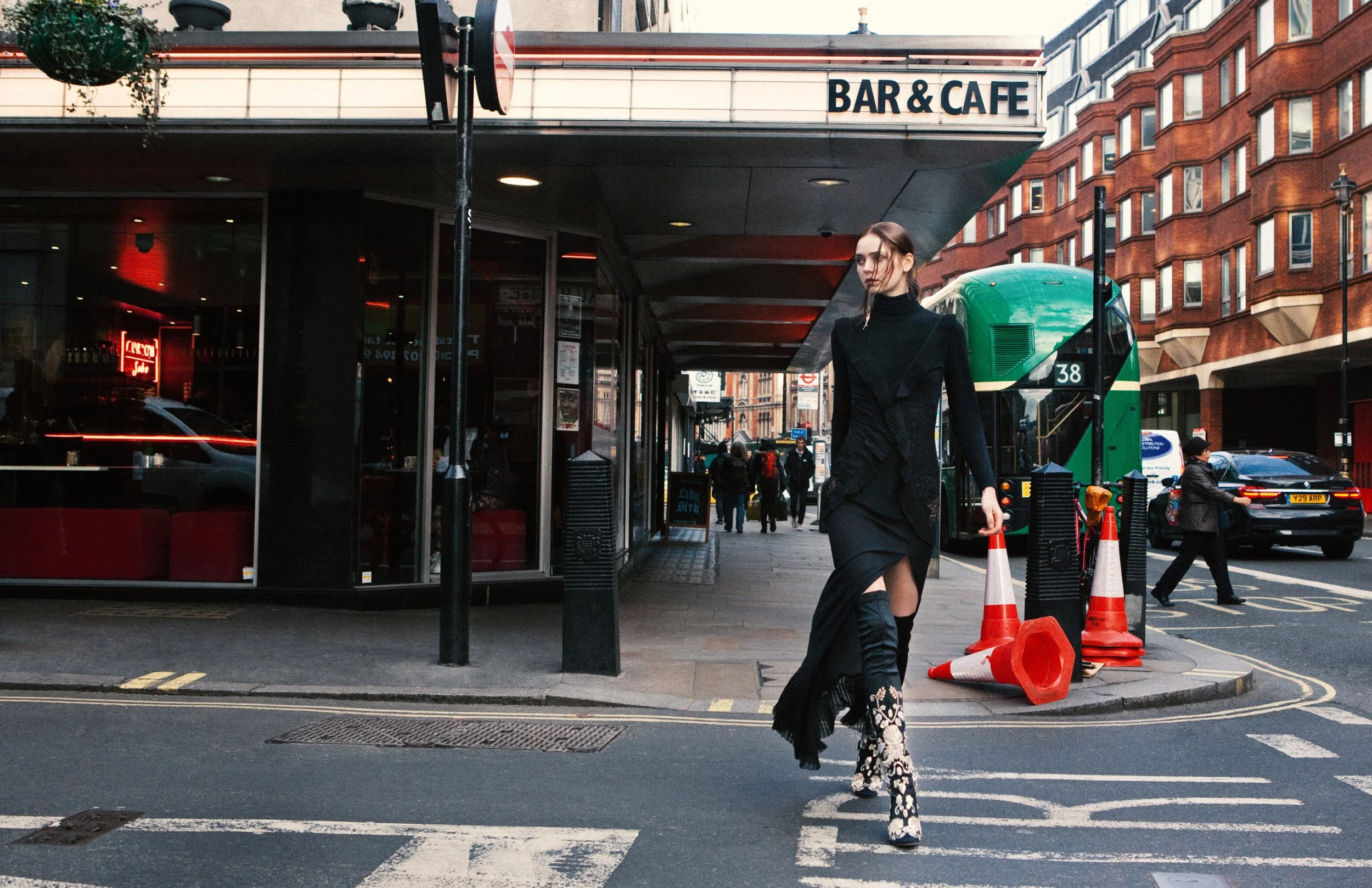 A woman in a black outfit walking across a city crosswalk in front of a bar and cafe, with a green bus and red brick buildings in the background, and orange traffic cones on the sidewalk.