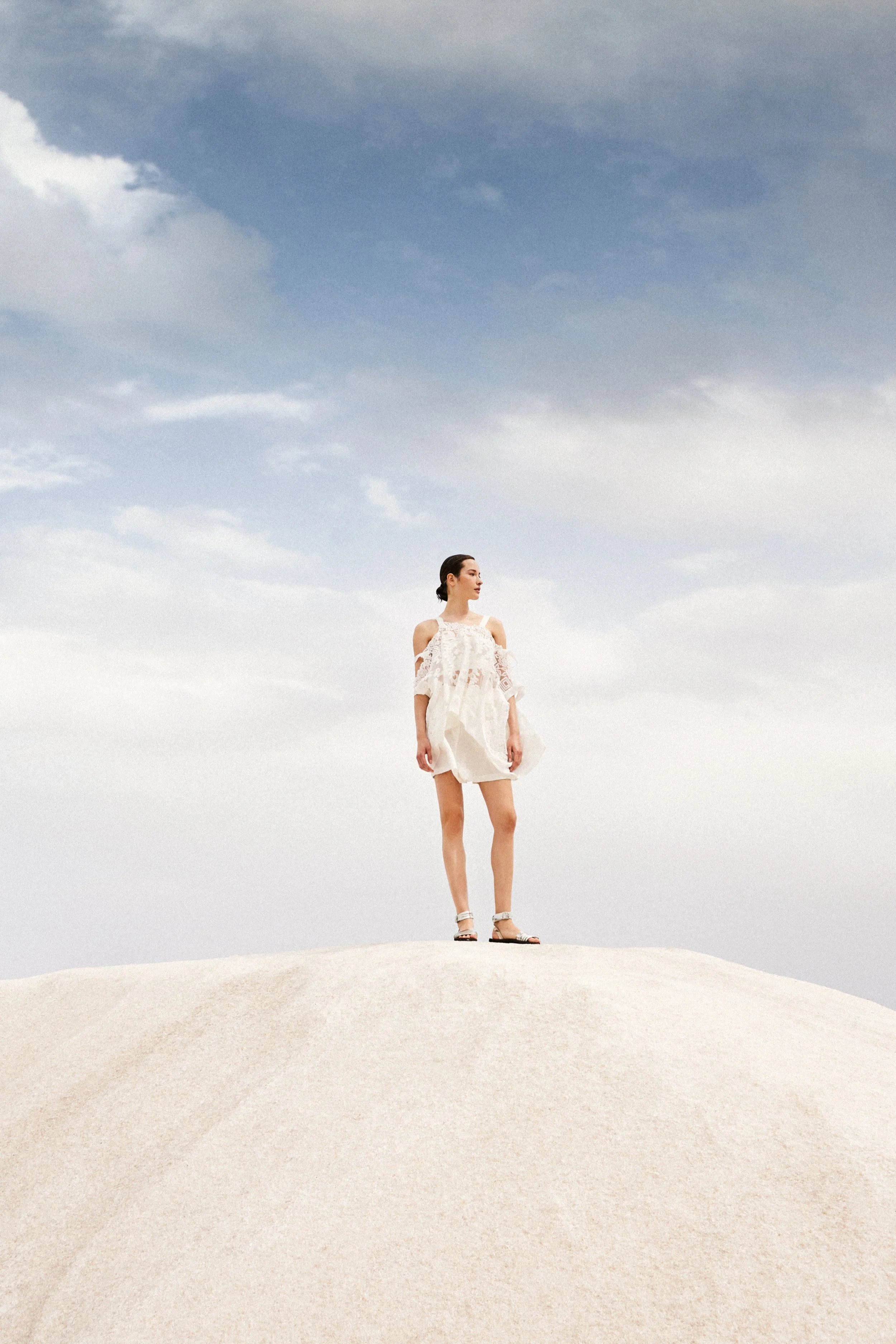 Woman in white dress and sandals standing on a white sandy hill under a partly cloudy sky.
