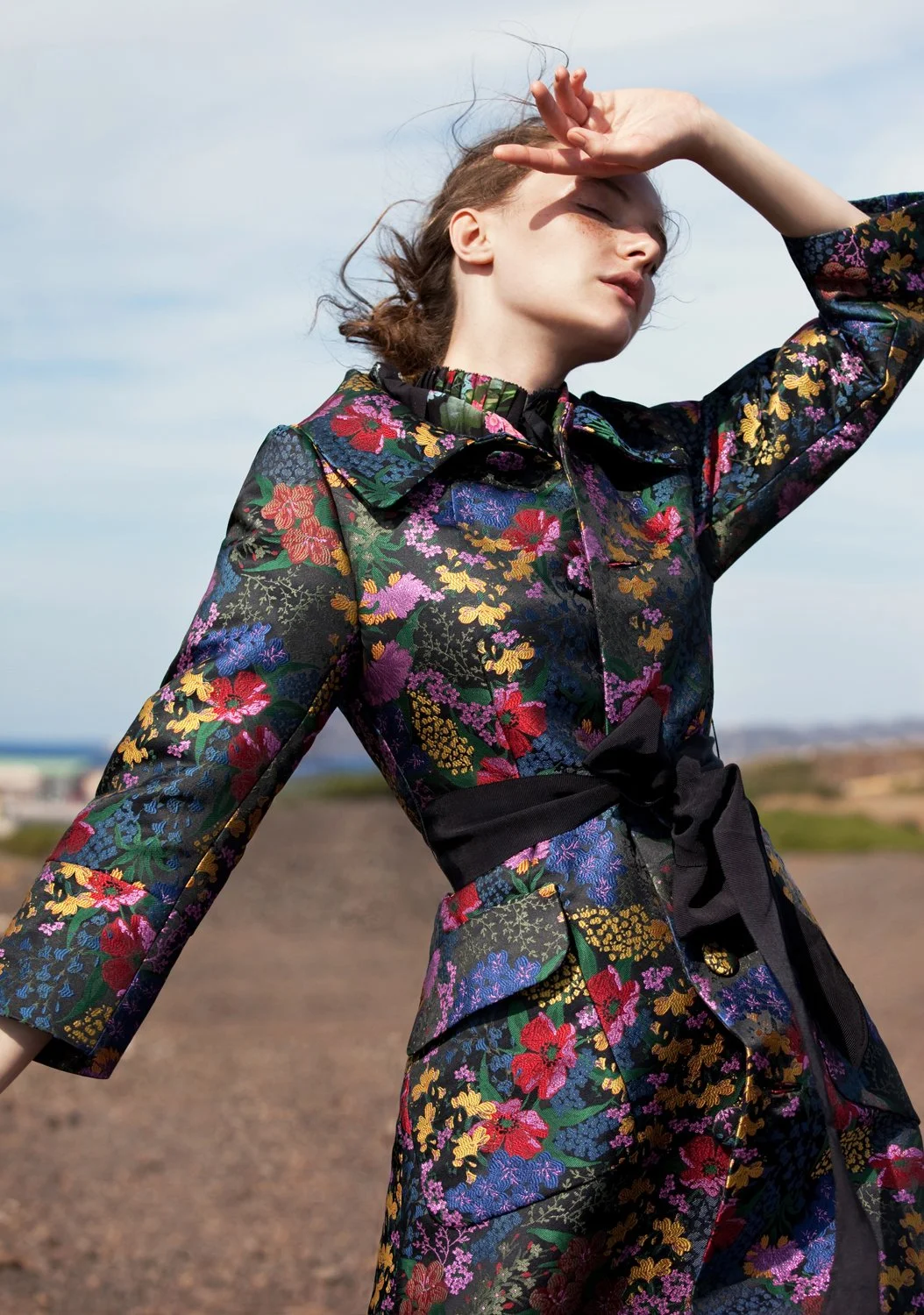 A woman with freckles and short hair shielding her eyes from the sun with her hand, wearing a floral patterned dress or jacket, standing outdoors in a rural or open landscape under a partly cloudy sky.
