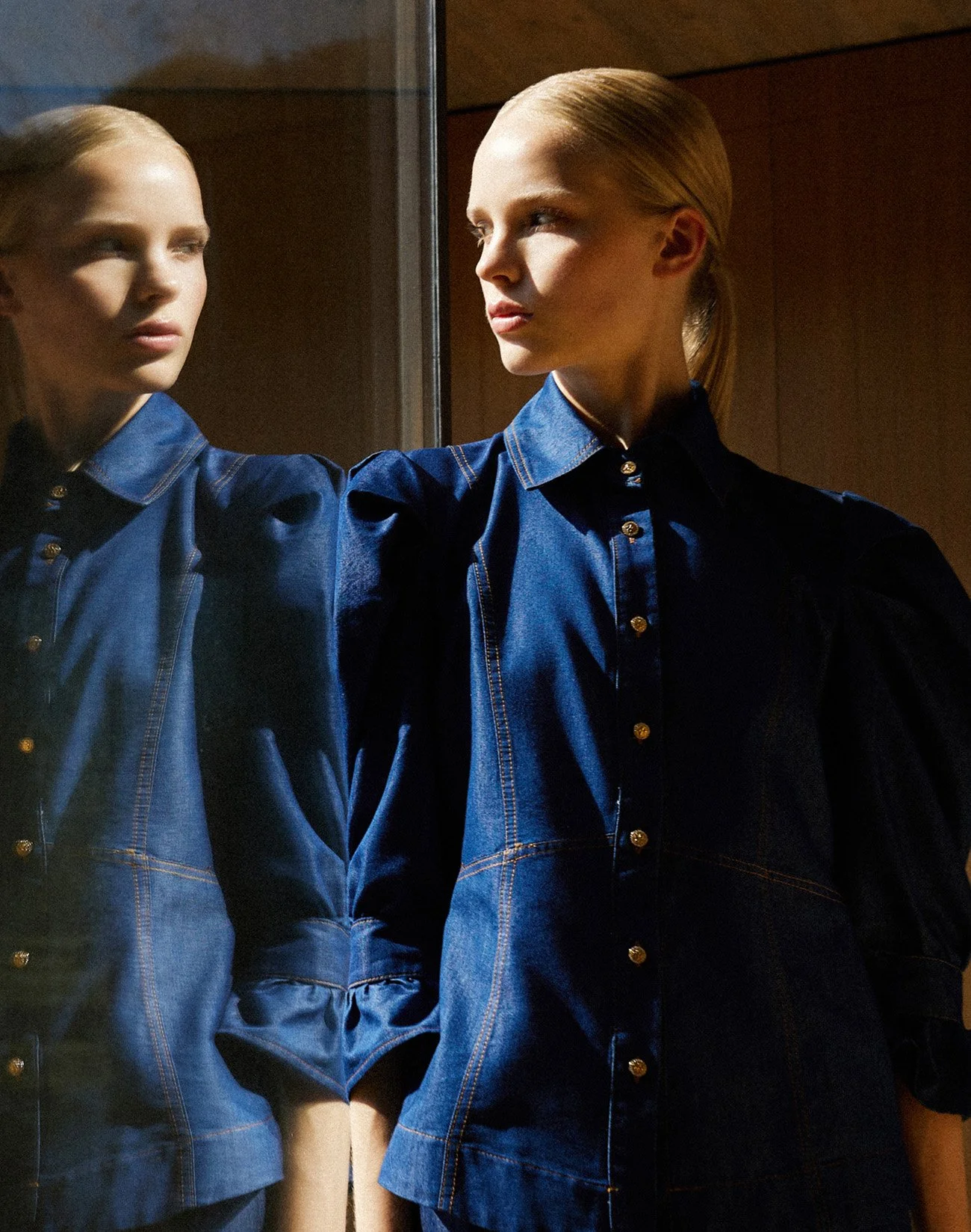 A woman wearing a blue denim shirt with gold buttons, standing next to a mirror, with her reflection visible.
