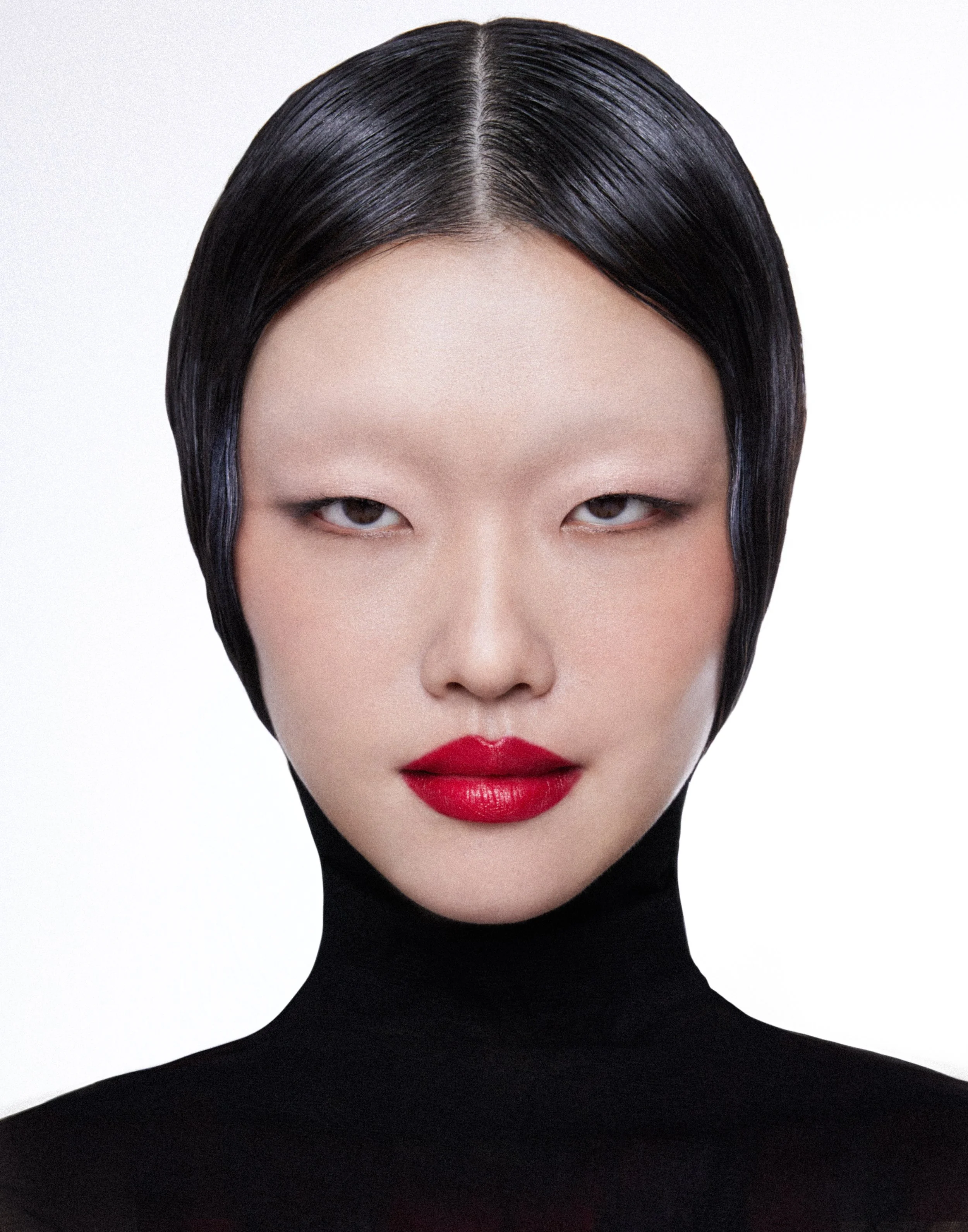 A woman with slicked-back black hair, wearing a black turtleneck, with bold red lipstick and minimal makeup, against a plain white background.