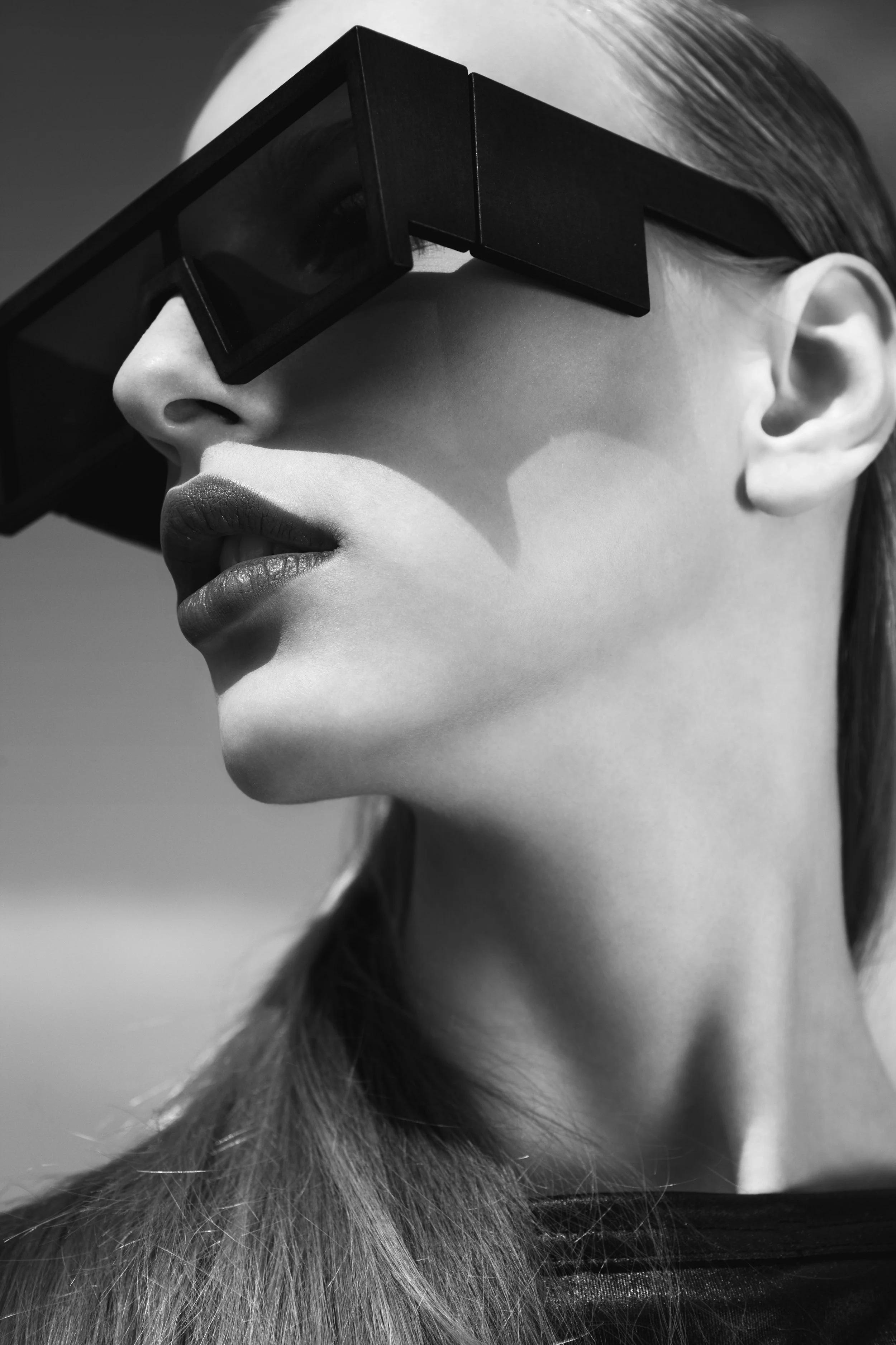 A close-up black and white photo of a woman wearing futuristic sunglasses, with her lips slightly parted and long hair flowing down.