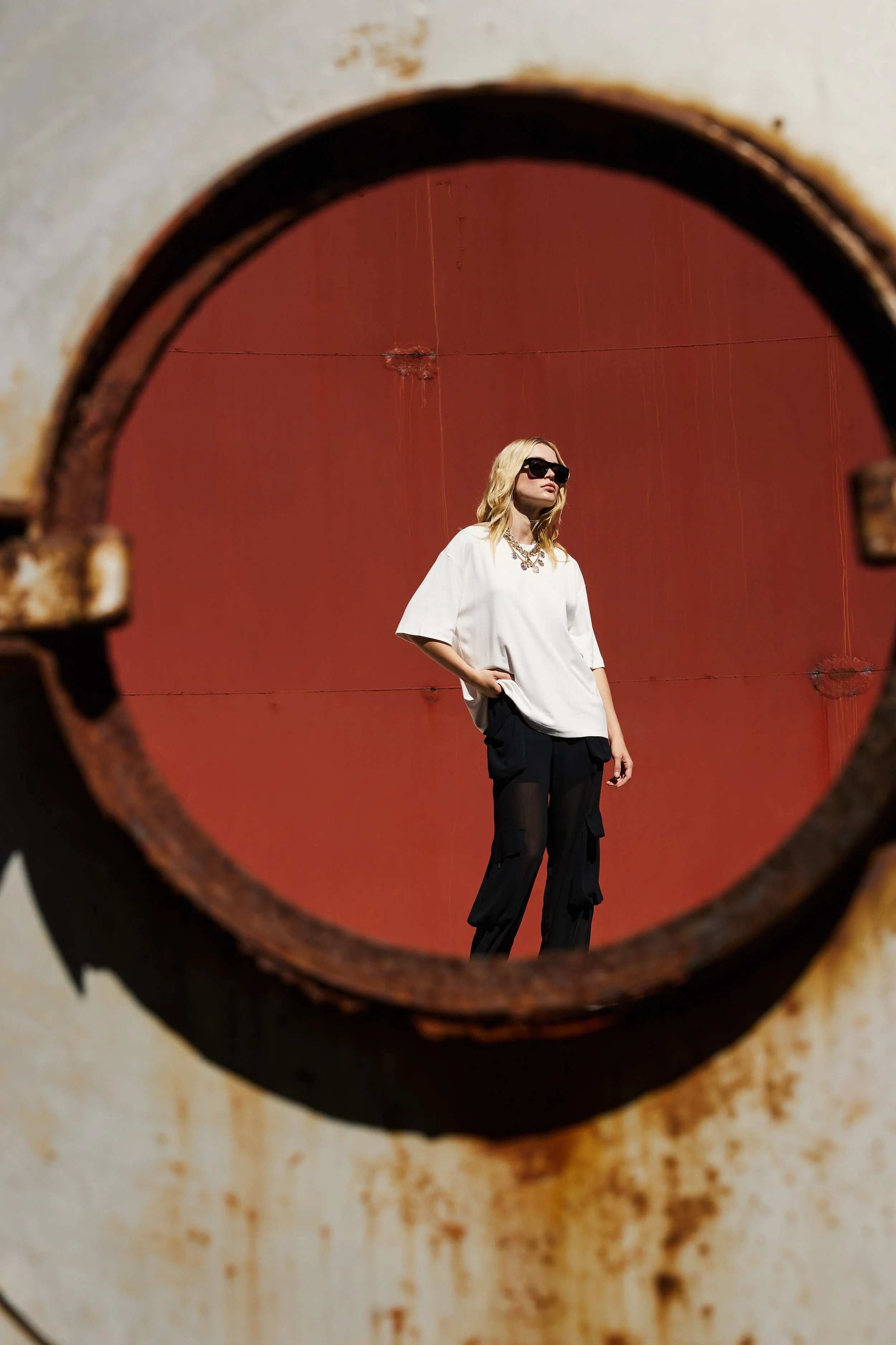 A woman wearing black sunglasses, a white oversized t-shirt, and black cargo pants stands against a red wall. The photo is taken through a rusty pipe opening, framing her silhouette.