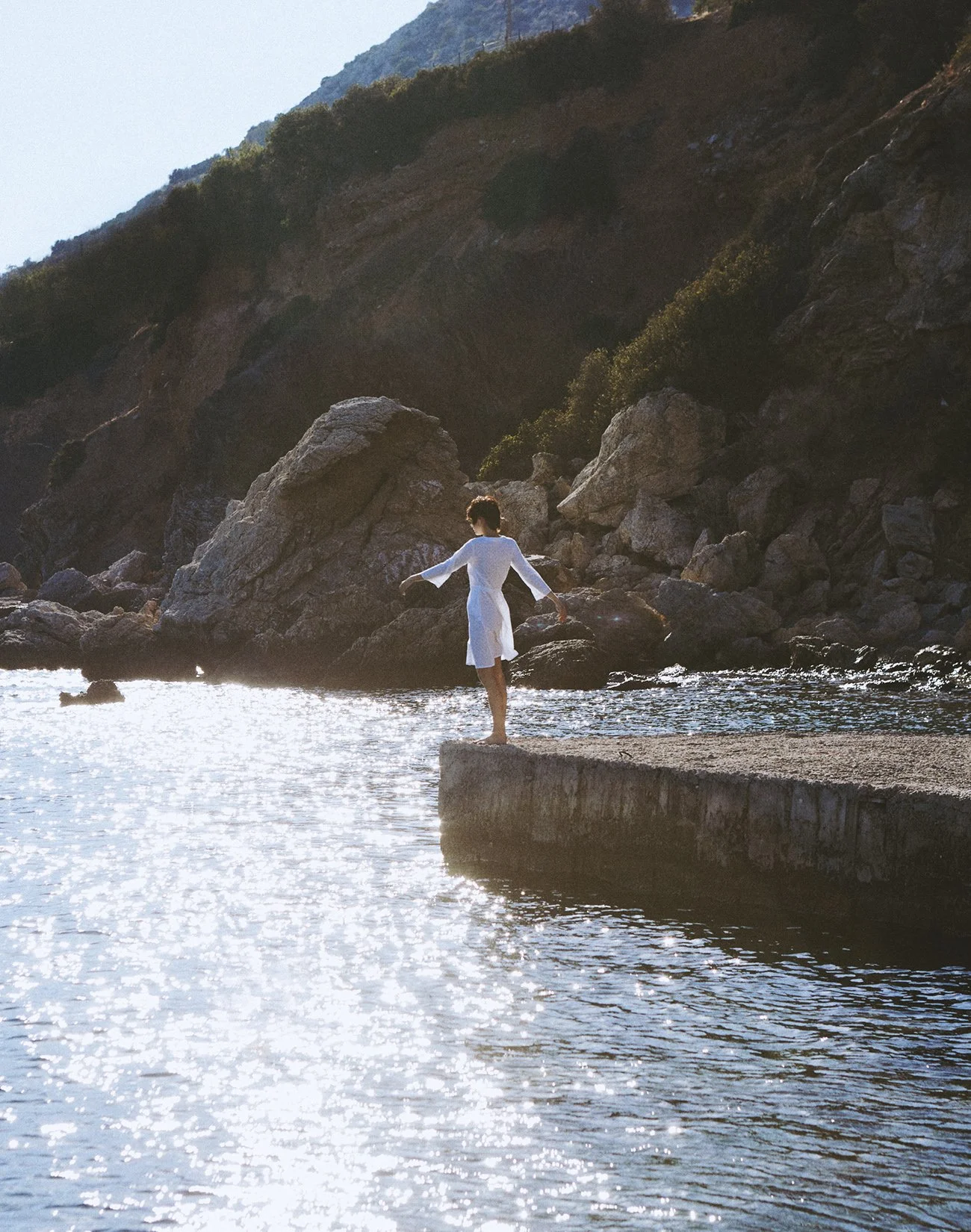 A woman in a white dress stands on a concrete dock at the edge of the water, balancing on one foot with her arms outstretched. She is near large rocks with a hillside covered in greenery in the background, and the sunlight reflects off the water.