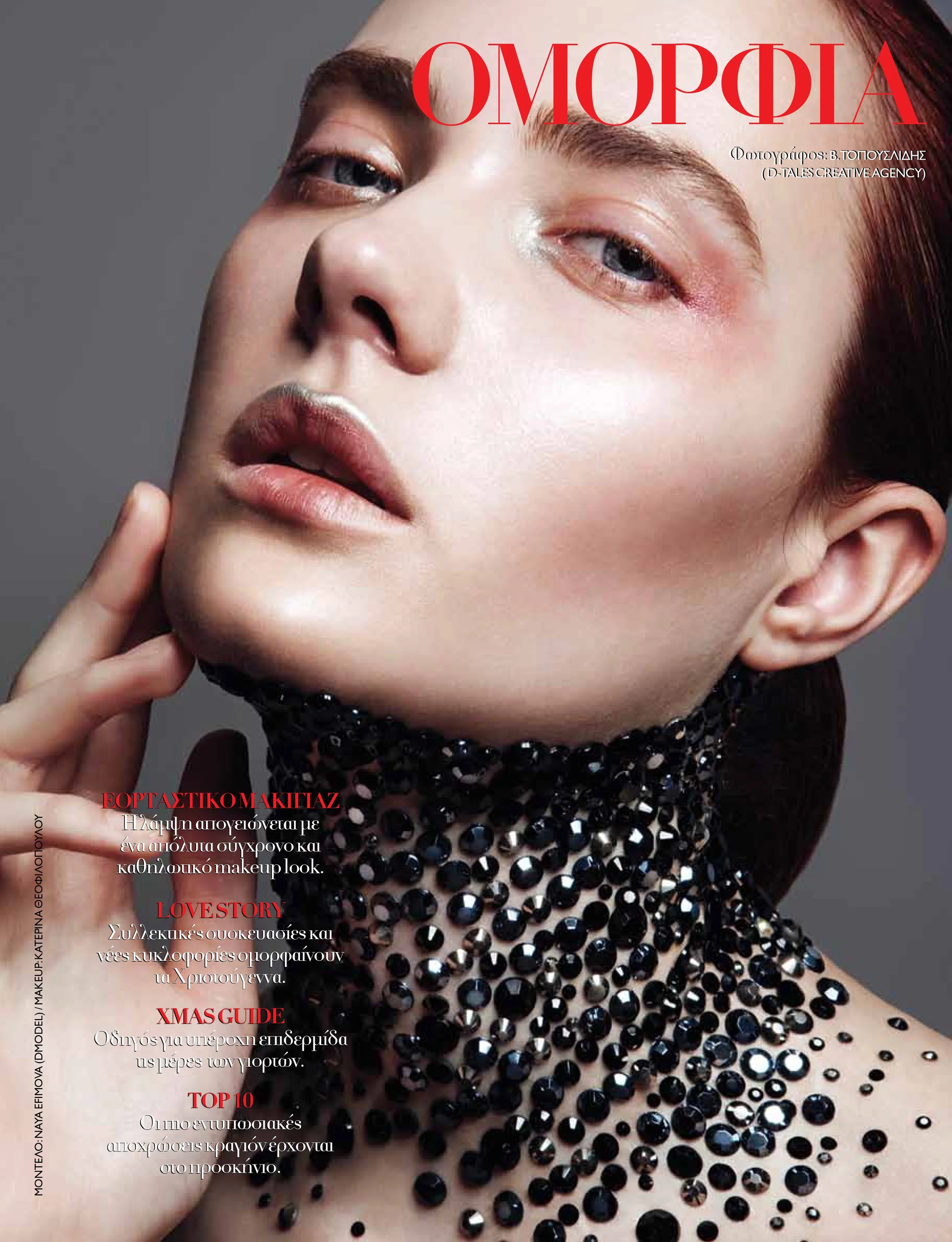 Close-up of a woman with pearl makeup, wearing a black sequin choker, touching her face with her hand, with red hair and makeup emphasizing her eyes and lips, on a gray background.