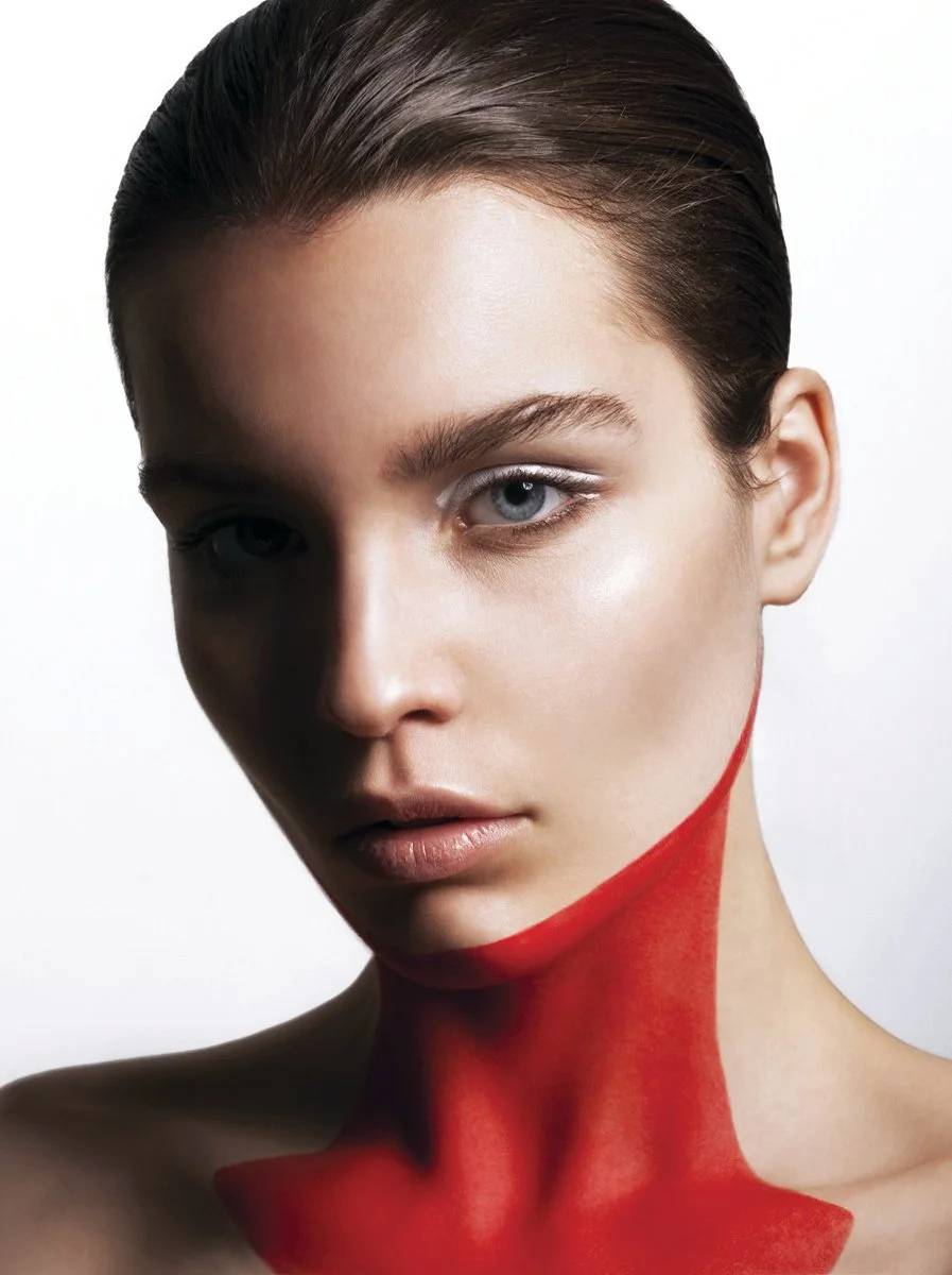 Close-up of a young woman with slicked-back brown hair and blue eyes, with red paint on her neck and shoulder.