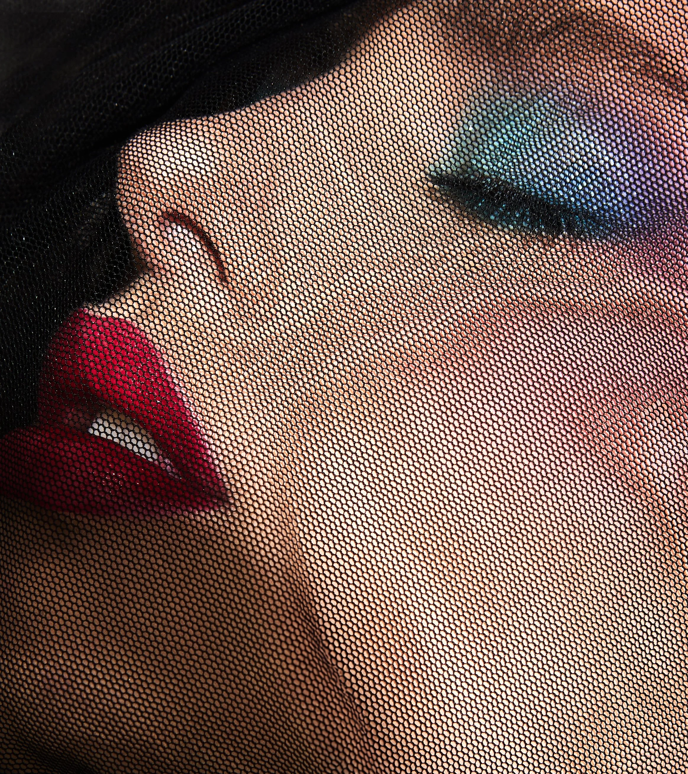Close-up of a woman's face behind a black netted fabric, showing her makeup with bold eyeshadow, eyeliner, and red lipstick.