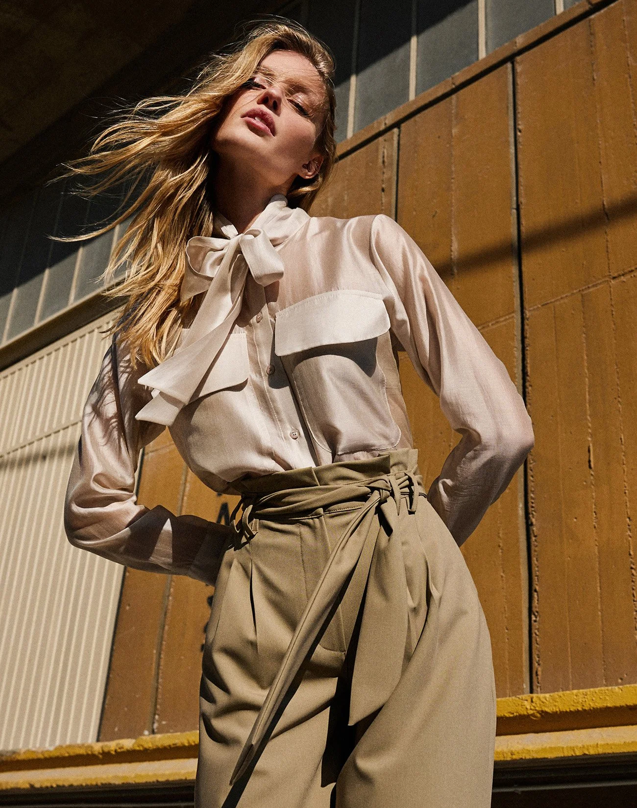 A woman wearing a cream-colored blouse with a bow tie and high-waisted beige pants standing outdoors, with a wooden wall and stairs in the background.
