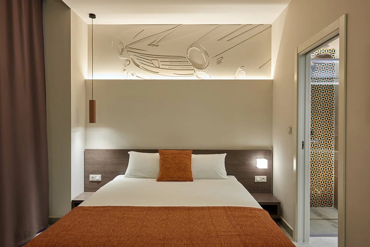 Chambre d'hôtel moderne avec lit, deux oreillers blancs, un coussin orange, lampe de nuit, mural avec design de voiture stylisée et porte donnant sur une salle de bain avec carrelage coloré.
