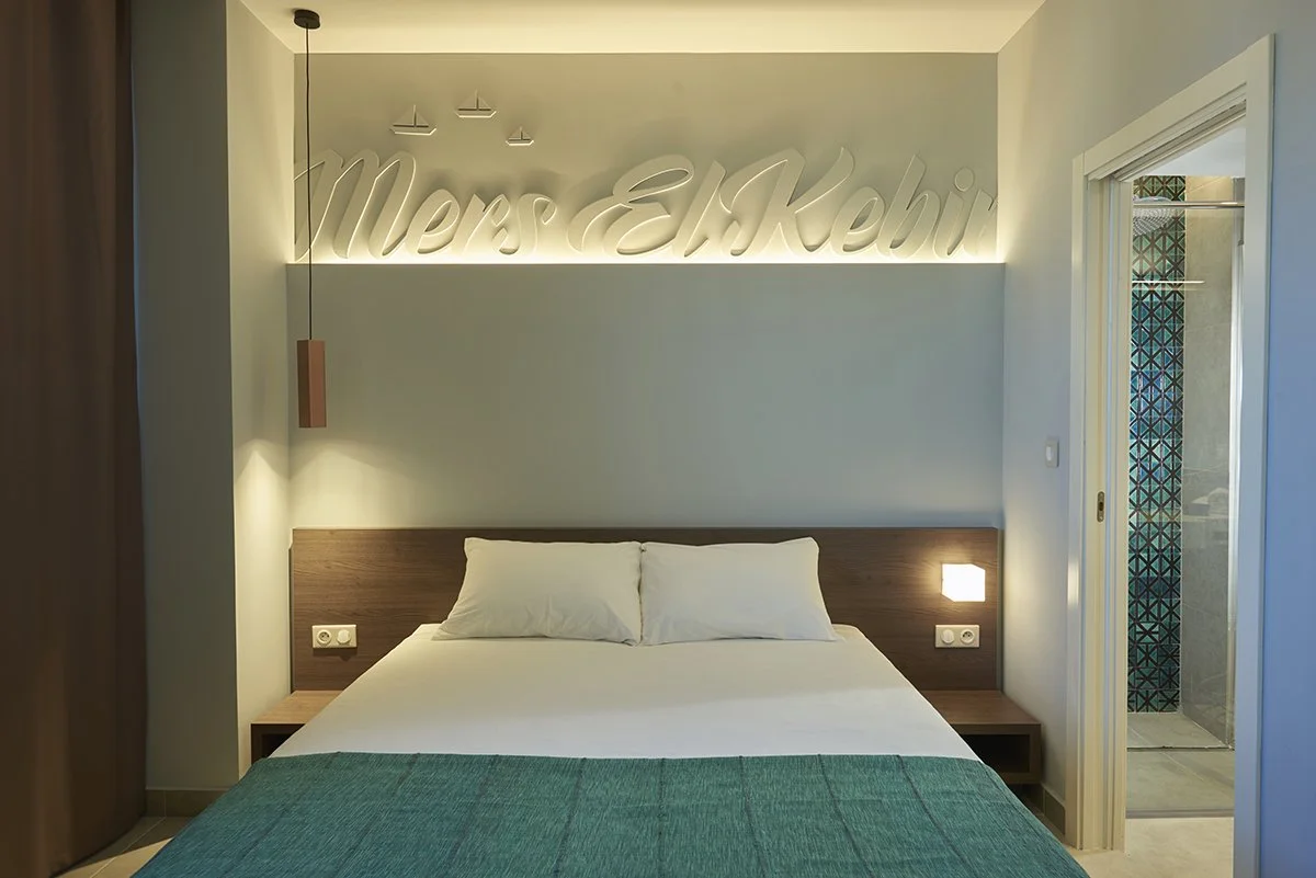 Chambre d'hôtel avec lit double, tête de lit en bois, deux lampes murales, inscription lumineuse 'Merci à Kehi' en arrière-plan, porte ouverte sur une salle de bain avec carrelage bleu et motifs géométriques.