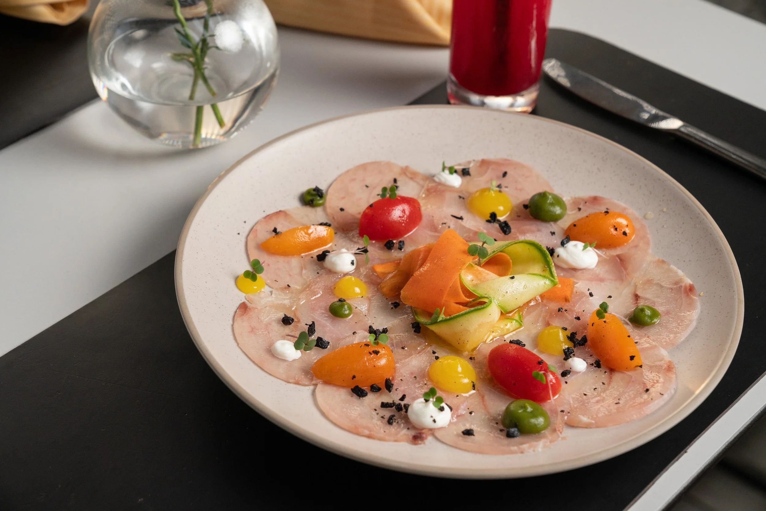 Assiette de carpaccio de viande garnie de petites tomates, de petites râpées de légumes, de micro-pousses, et de petites sauces blanches. En arrière-plan, un verre d'eau avec une fleur, un verre de jus rouge, et des couverts.