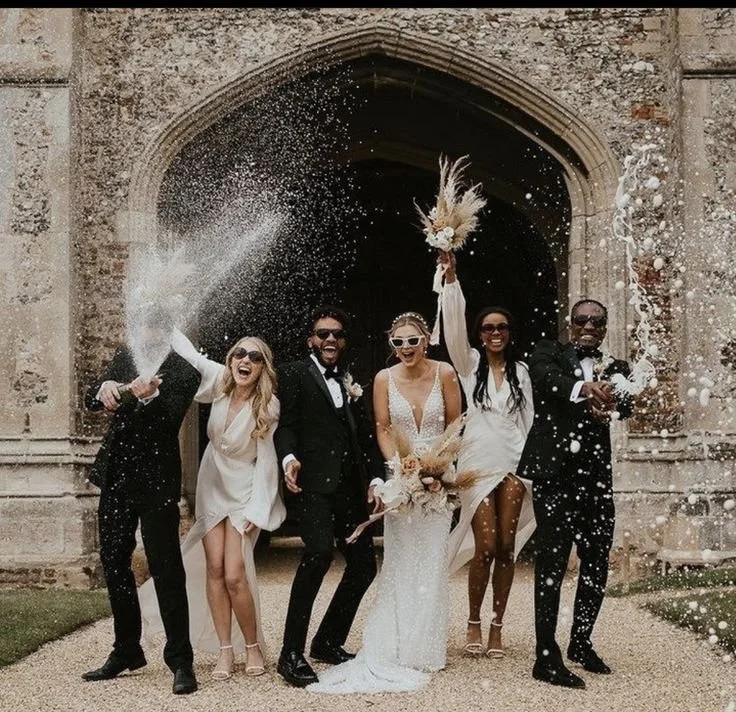 Un groupe de personnes en costumes de mariage célébrant devant une vieille porte en pierre, avec des confettis en l'air.