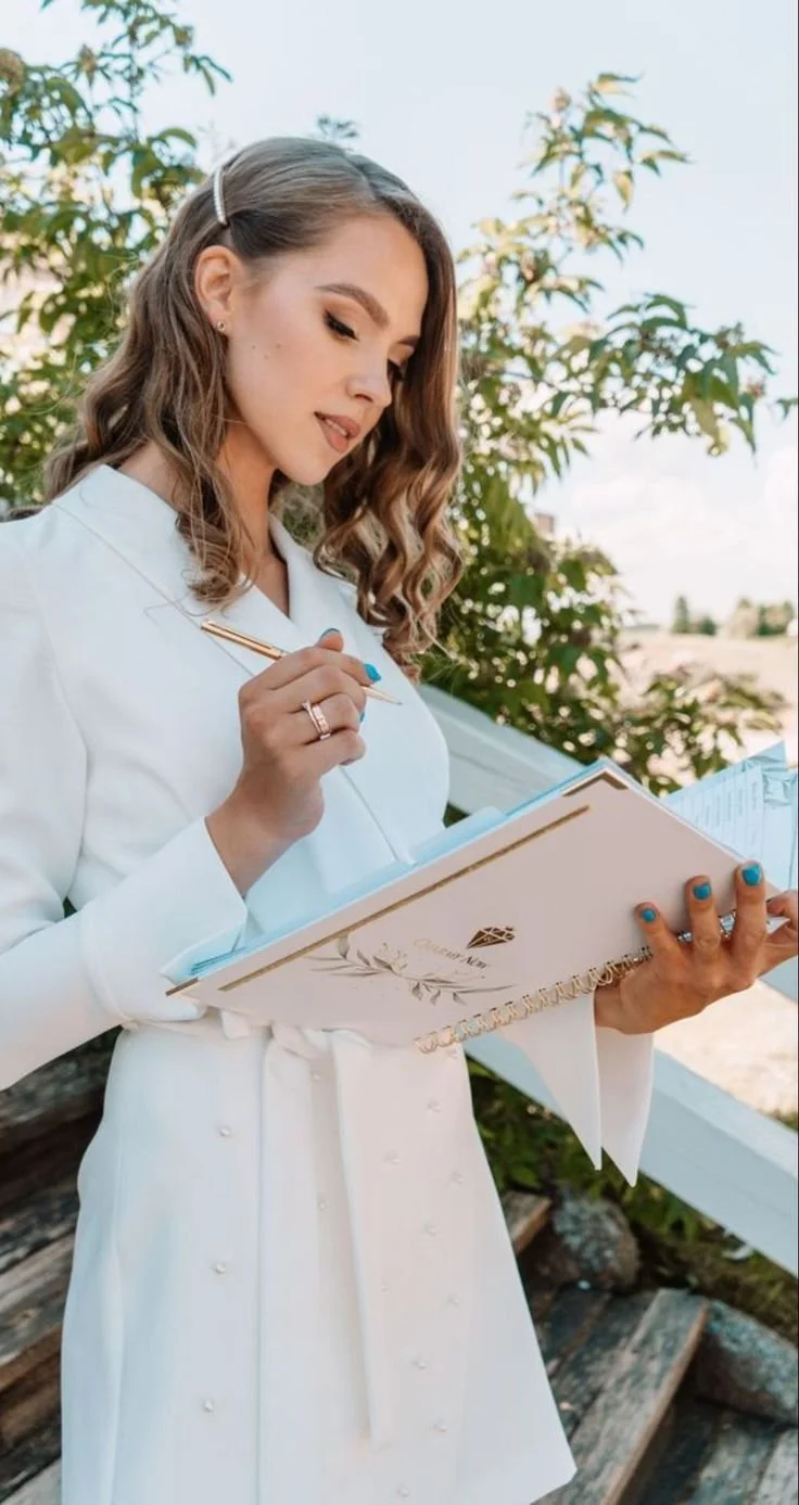 Une femme en robe blanche écrit dans un cahier dehors, avec des arbres et un ciel clair en arrière-plan.