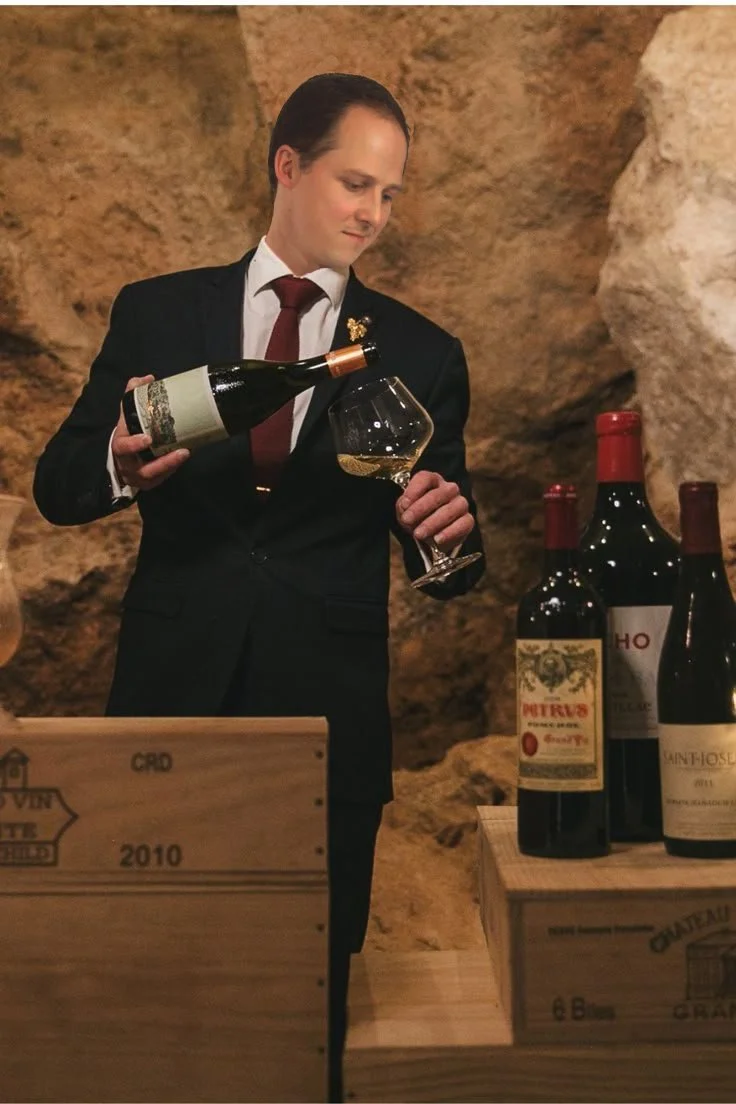 Un homme en costume noir verse du vin dans un verre lors d'une dégustation de vins, avec plusieurs bouteilles sur une table en bois dans un cave à vin.