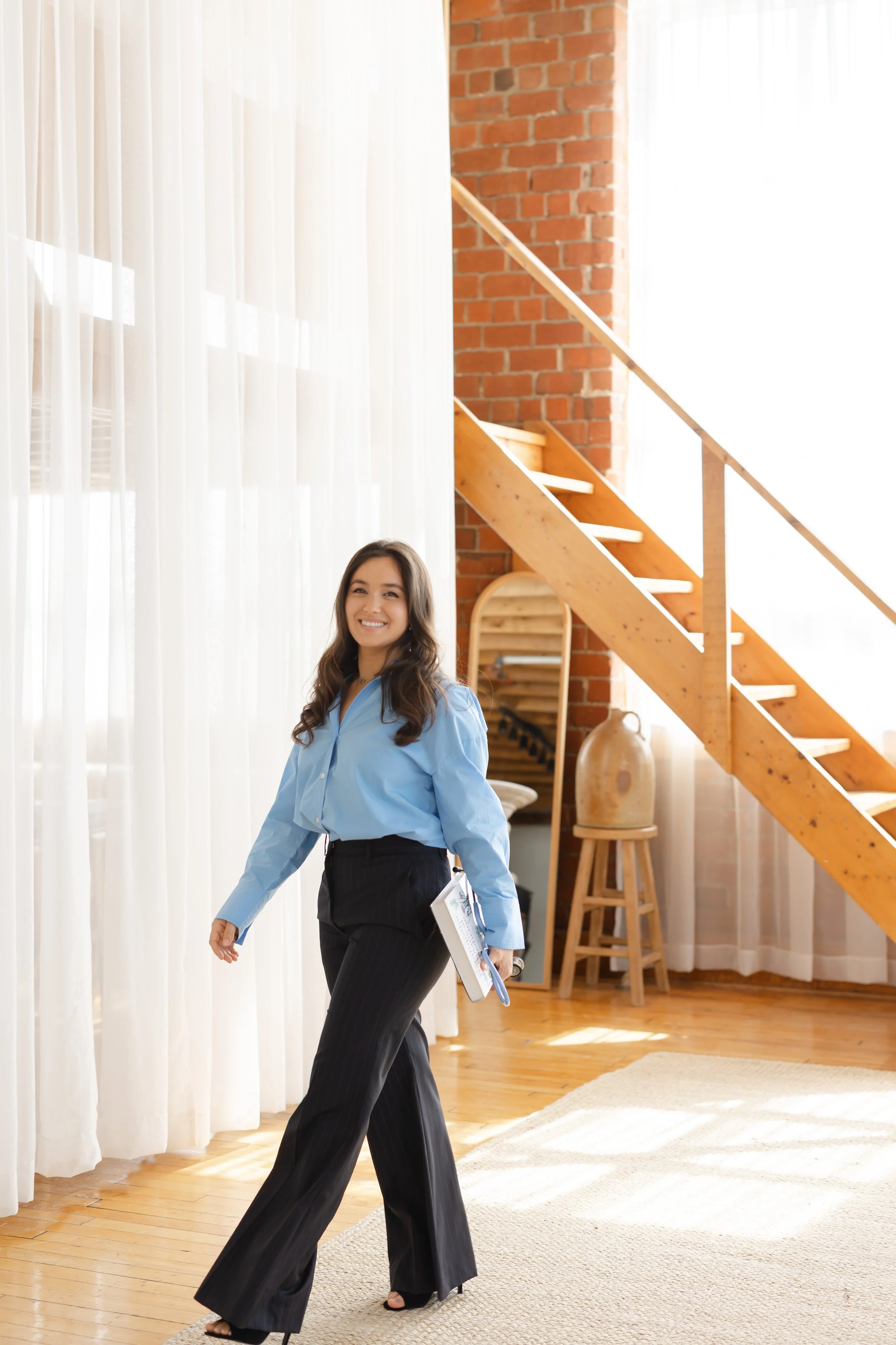 A young woman with long dark hair wearing a light blue blouse and dark pinstripe pants walking inside a well-lit room with a staircase and brick wall in the background, smiling and carrying a clipboard or notebook.