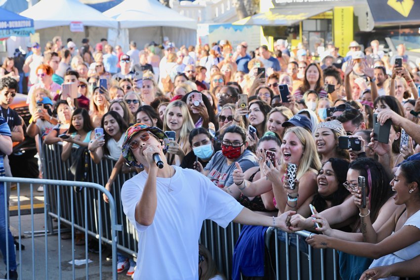 A female singer performs at an outdoor concert surrounded by a large, enthusiastic crowd of fans taking photos and videos.