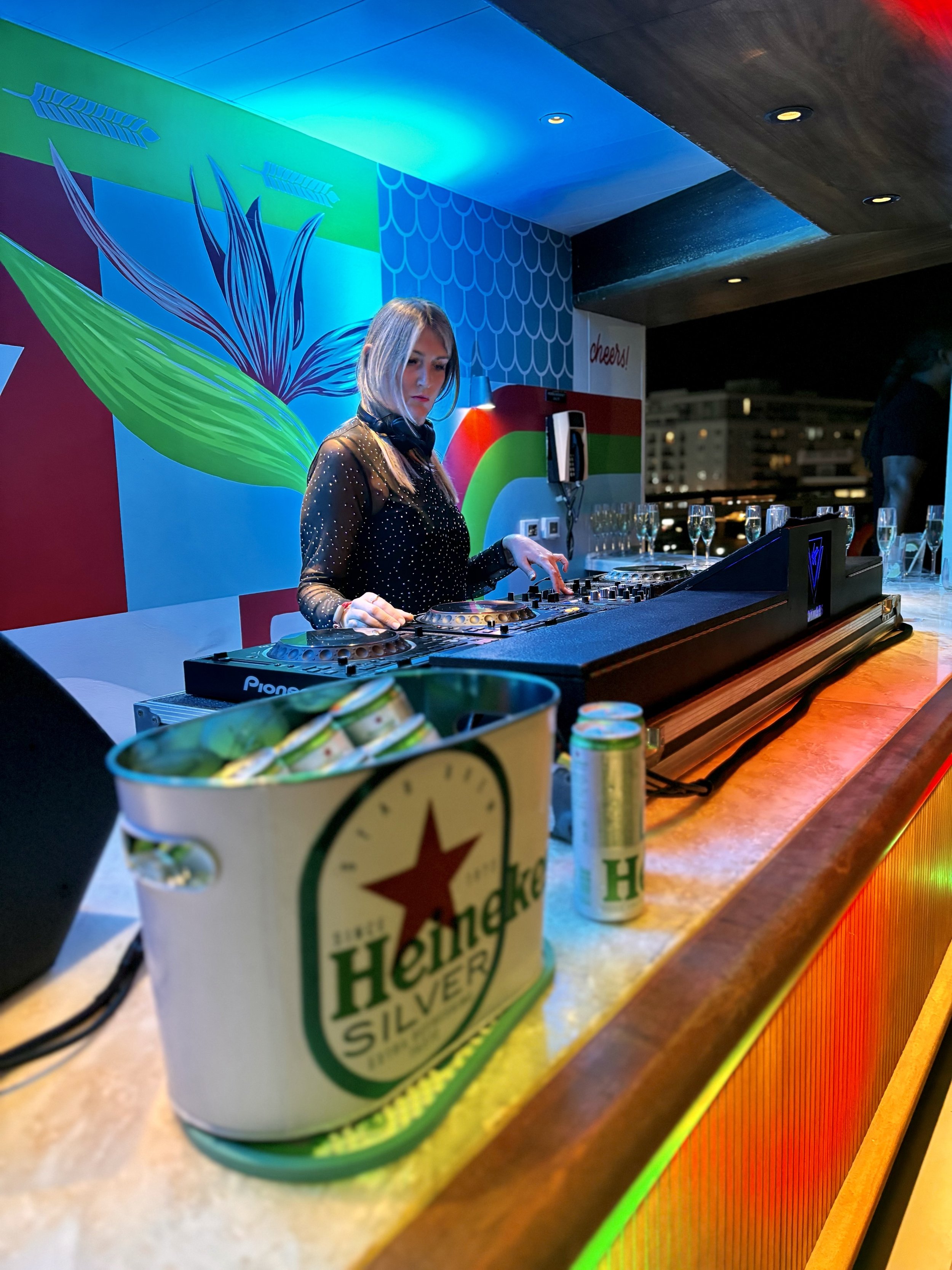 A female DJ with blonde hair, wearing a black dress with sheer sleeves, playing music on DJ equipment at a bar or club. There is a bucket of Heineken Silver cans and a few cans on the bar counter. The background features colorful wall art with geomet