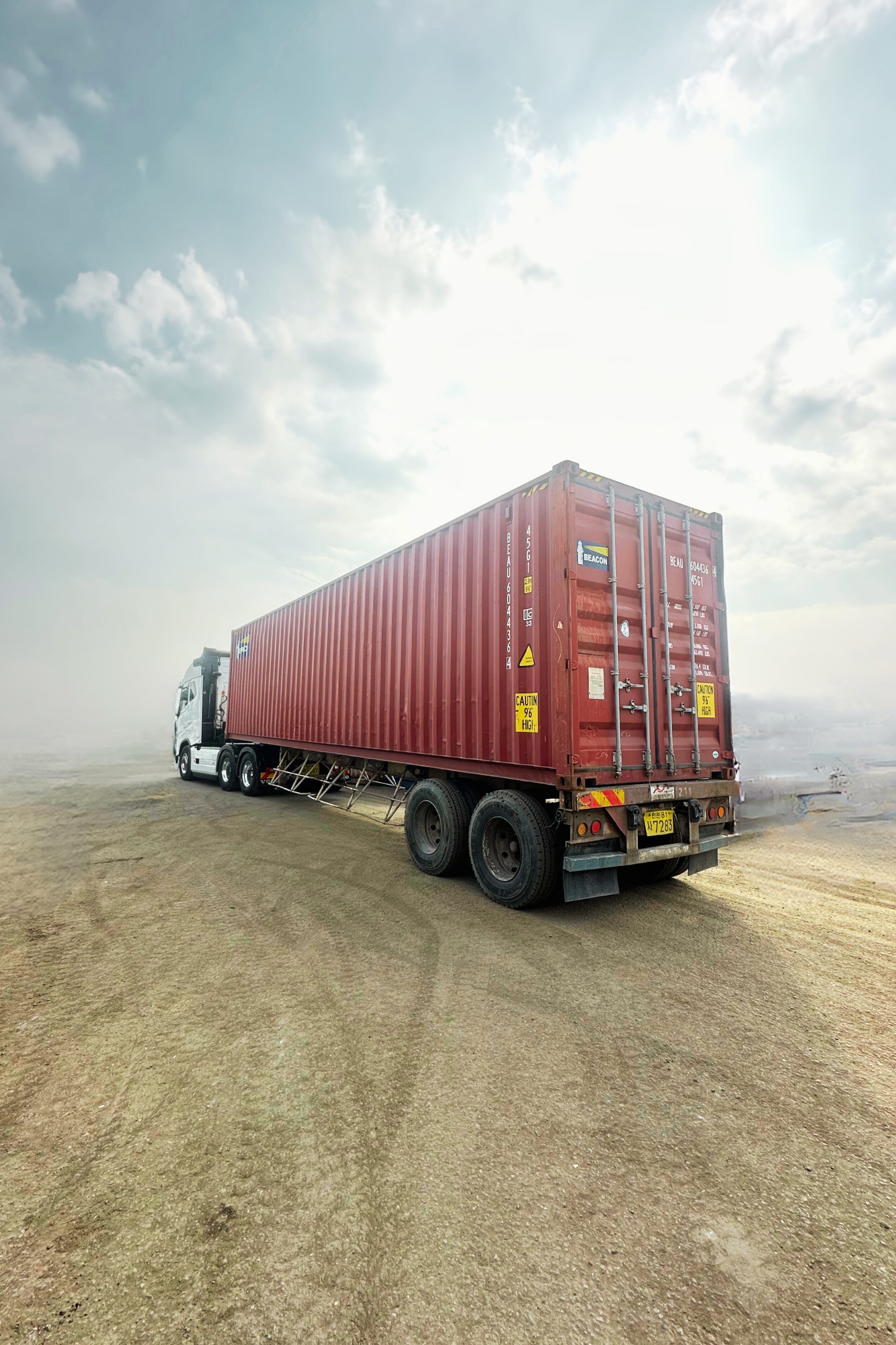A semi-truck with a red shipping container on a dirt surface under a cloudy sky.