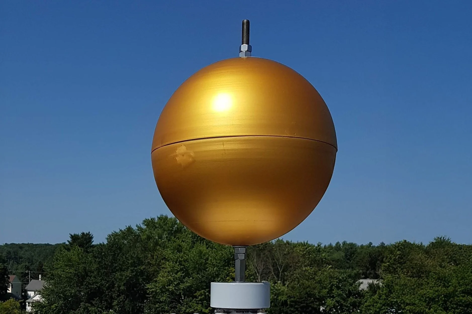 Finial Ball, mounted on a cell tower against a clear blue sky with green trees in the background.