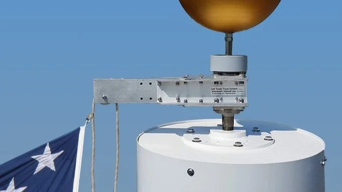 The top of a cell tower with an Adapter Plate, SwingArm Truck, Finial Ball, Halyard, and American Flag against a blue sky.