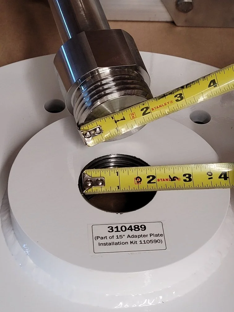 A metal pipe fitting on a white circular Adapter Plate with measurement tapes showing approximately 2.5 inches in diameter.