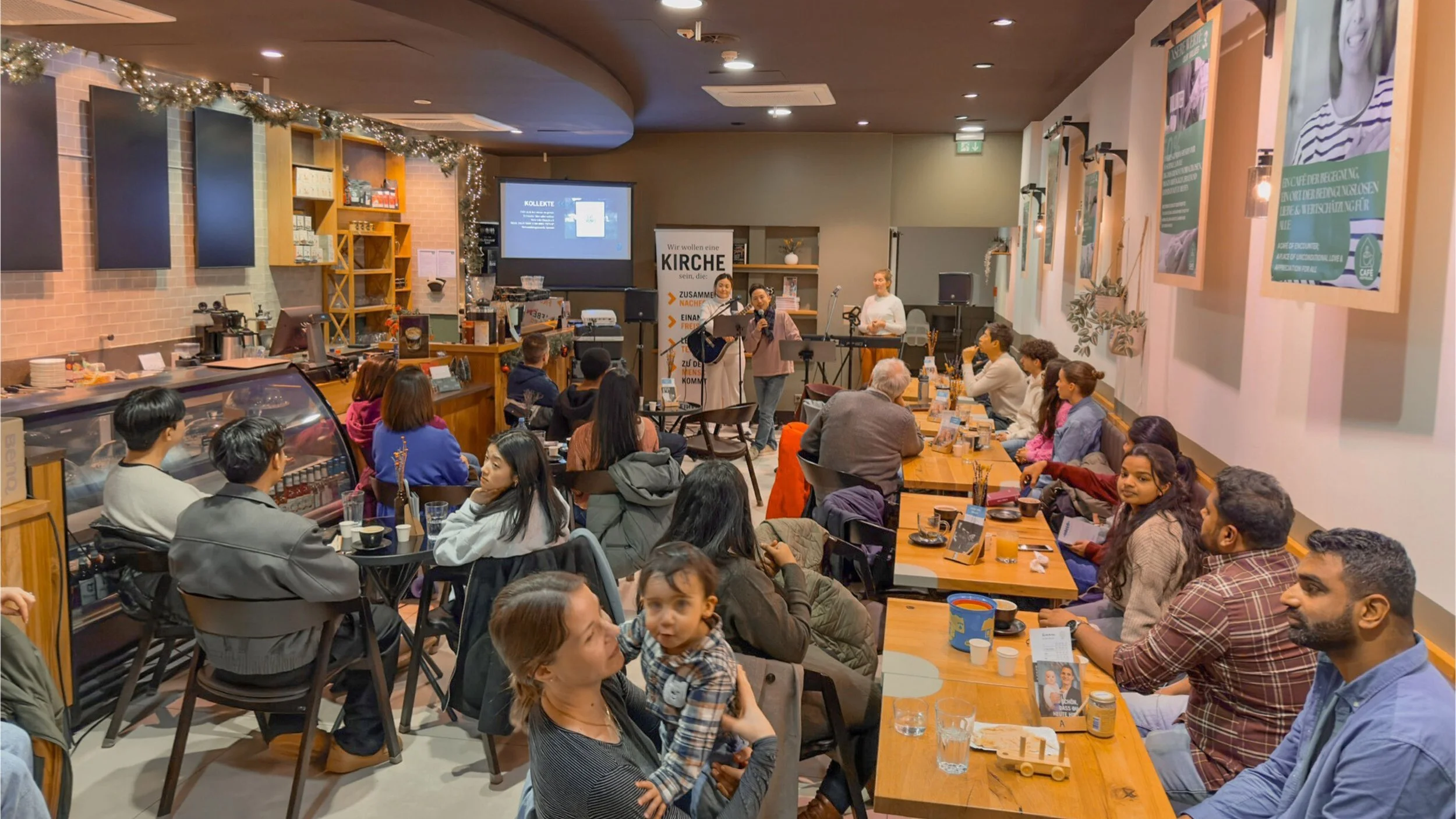 Menschen sitzen bei einem Gottesdienst in einem Café, im Vordergrund hält eine Frau ein kleines Kind und lächelt in die Kamera. Im Hintergrund singen zwei Musiker auf einer kleinen Bühne.