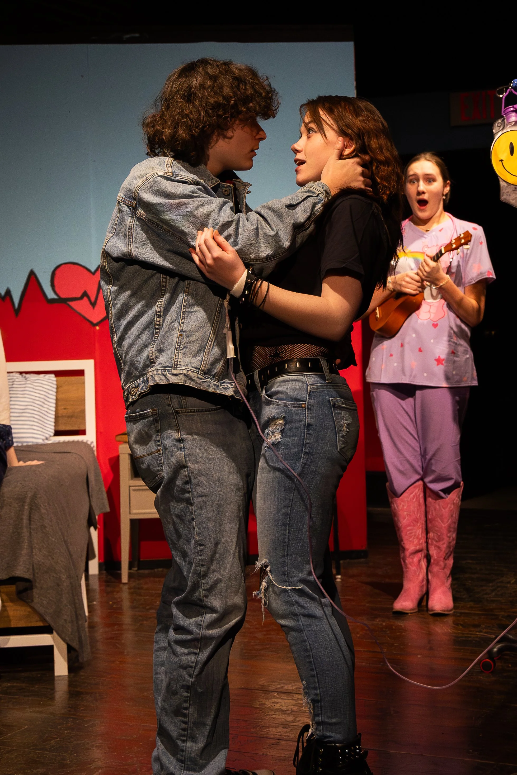 A boy and a girl in an embrace with a shocked girl in purple scrubs holding a ukulele.