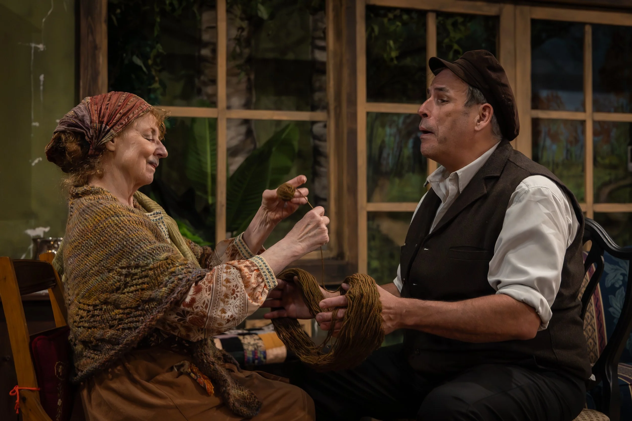 Lady in a scarf and shawl rolling a ball of yarn while a man I a vest and hold fashioned hat holds the yarn on his hand for her. 