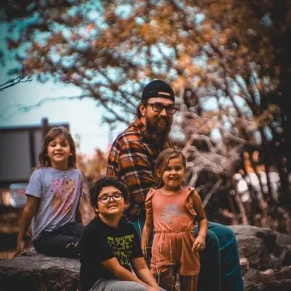 A man with glasses and a beard sitting outdoors with three young girls, surrounded by autumn trees.