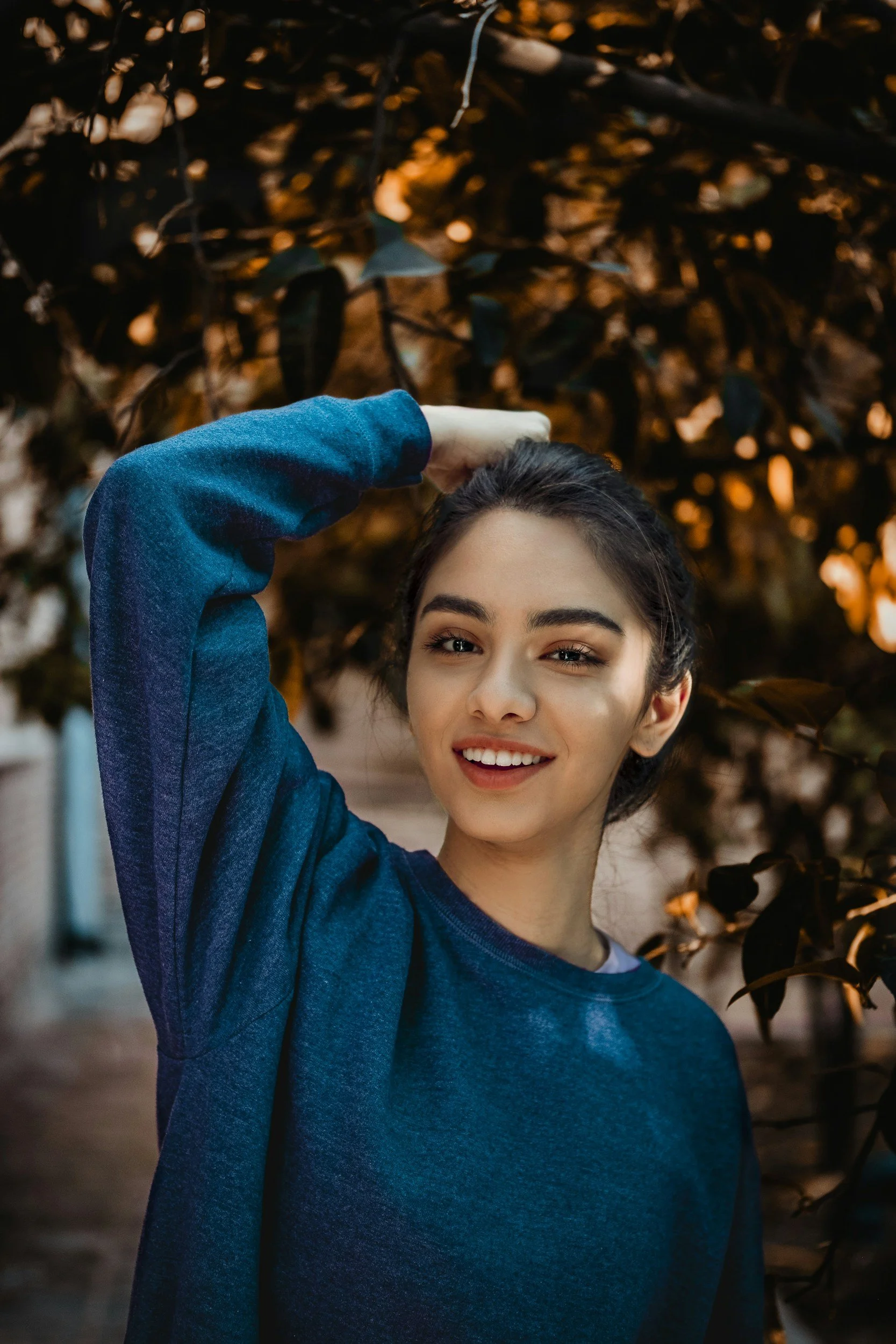 A young woman smiling and looking at the camera, wearing a blue sweatshirt, standing outdoors with blurred brown and orange leaves in the background. She has dark hair and eyebrows, and one hand is raised, touching her hair.