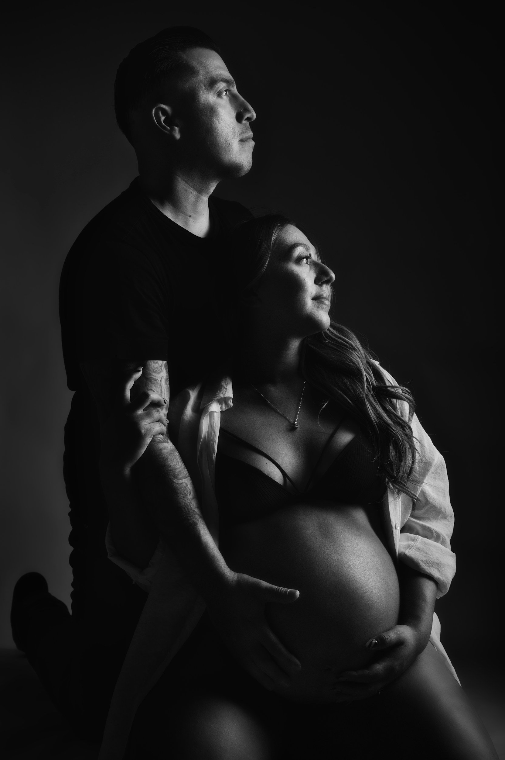 Black and white photograph of a man and woman in an intimate pose. The man stands behind the woman, looking upward, while the woman looks slightly upward with a smile. They are both partially dressed, with the woman's midriff exposed, and show affectionate body language.
