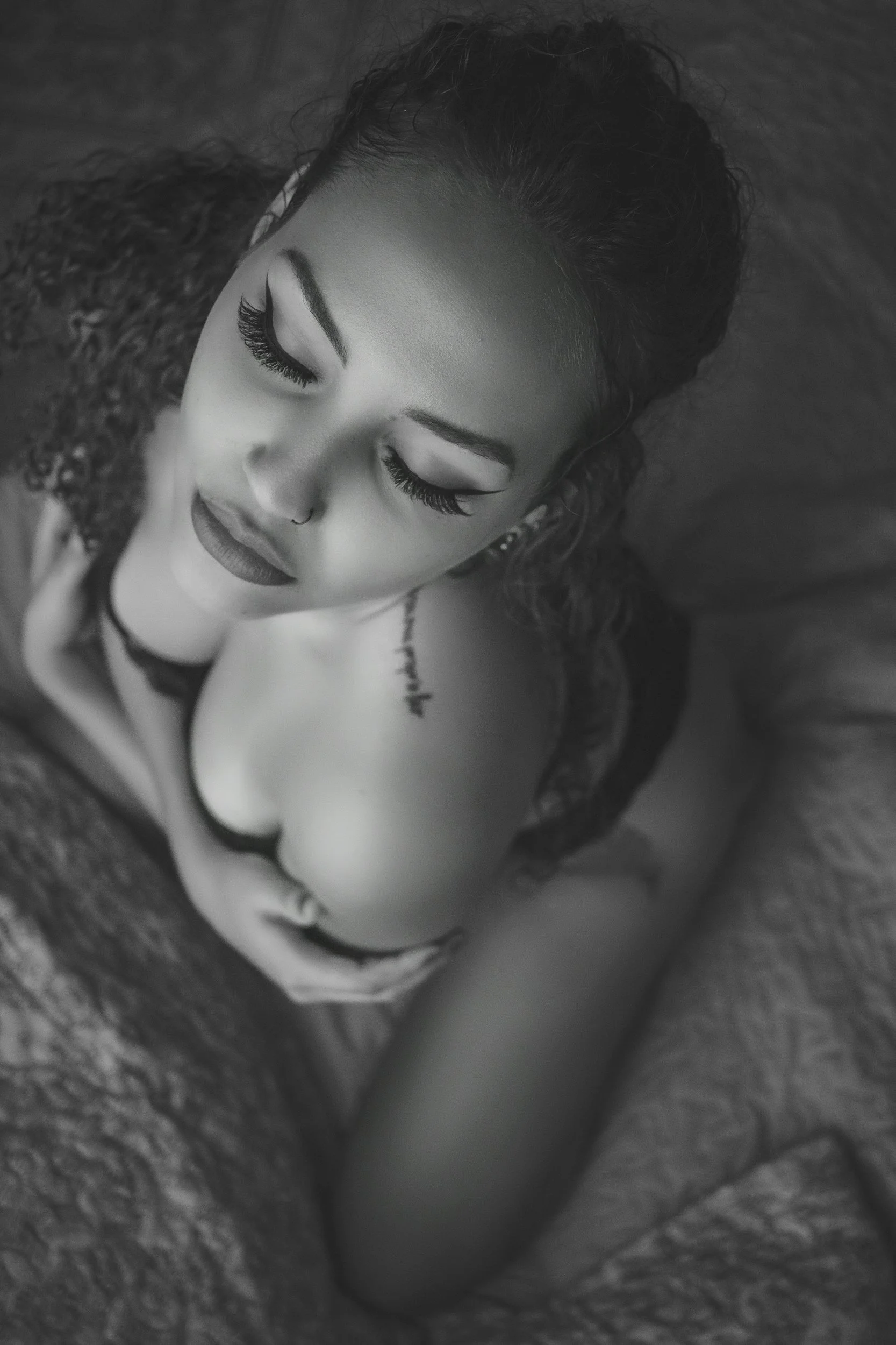 Close-up of a woman with curly hair, closed eyes, long eyelashes, and makeup, lying on a soft surface with her eyes closed.